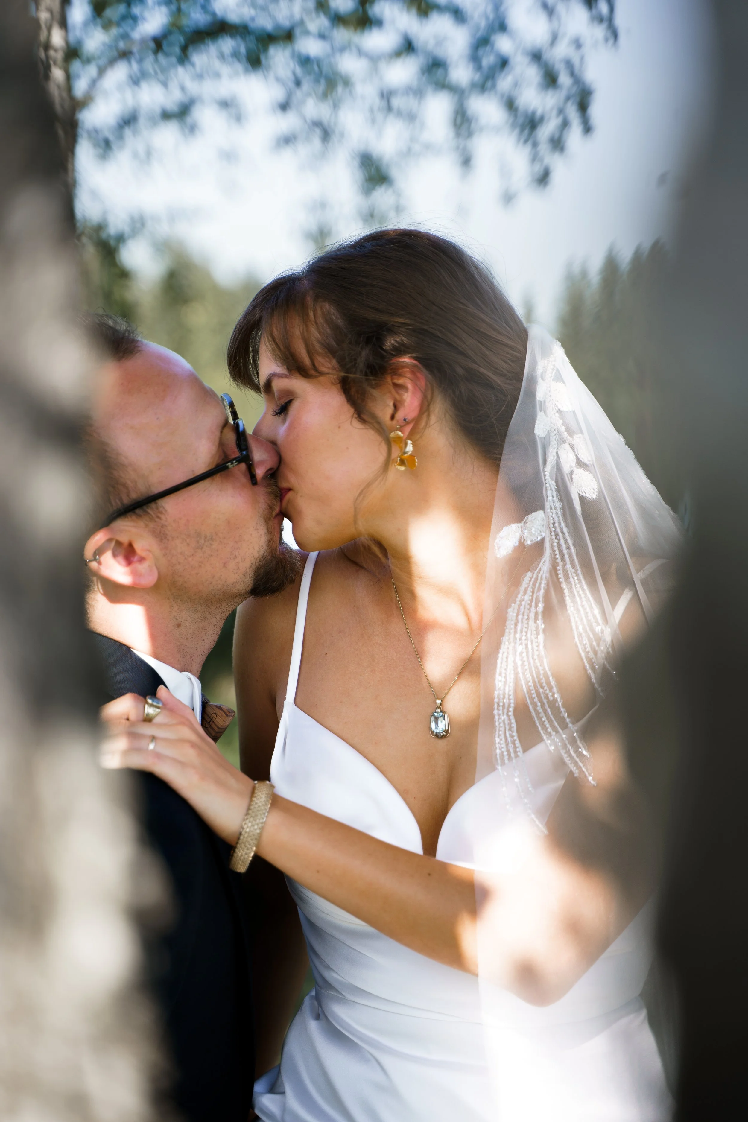 Ein Brautpaar küsst sich draußen, umgeben von Bäumen, bei Sonnenlicht, im Hochzeitskleid und Anzug.