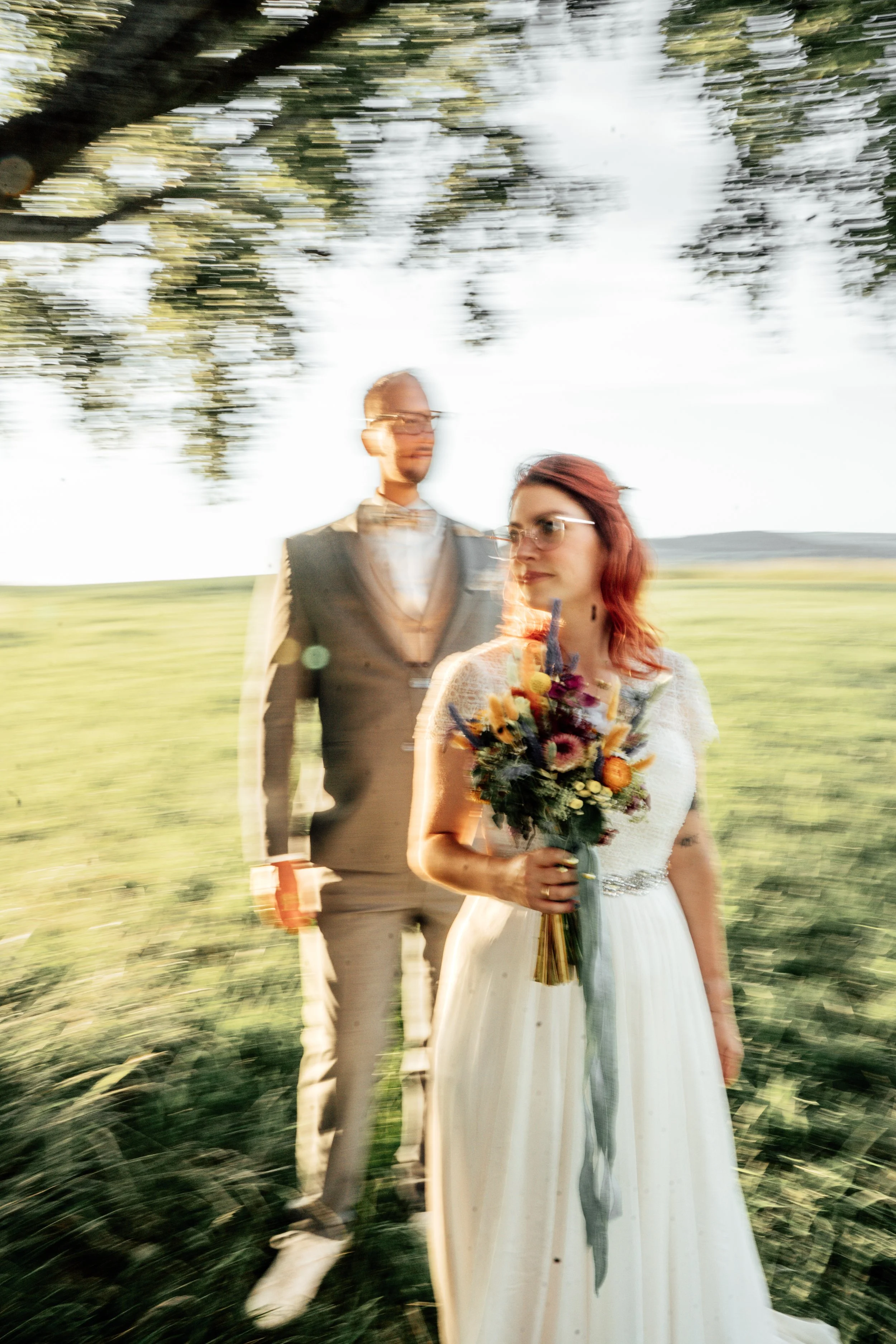 Ein Brautpaar steht im Freien, die Frau hält einen Blumenstrauß, im Hintergrund sind Bäume und eine weite Wiese, das Bild ist unscharf und wirkt Bewegung schräg. Das Paar trägt formelle Kleidung, sie scheint ein Hochzeitsfoto zu machen.
