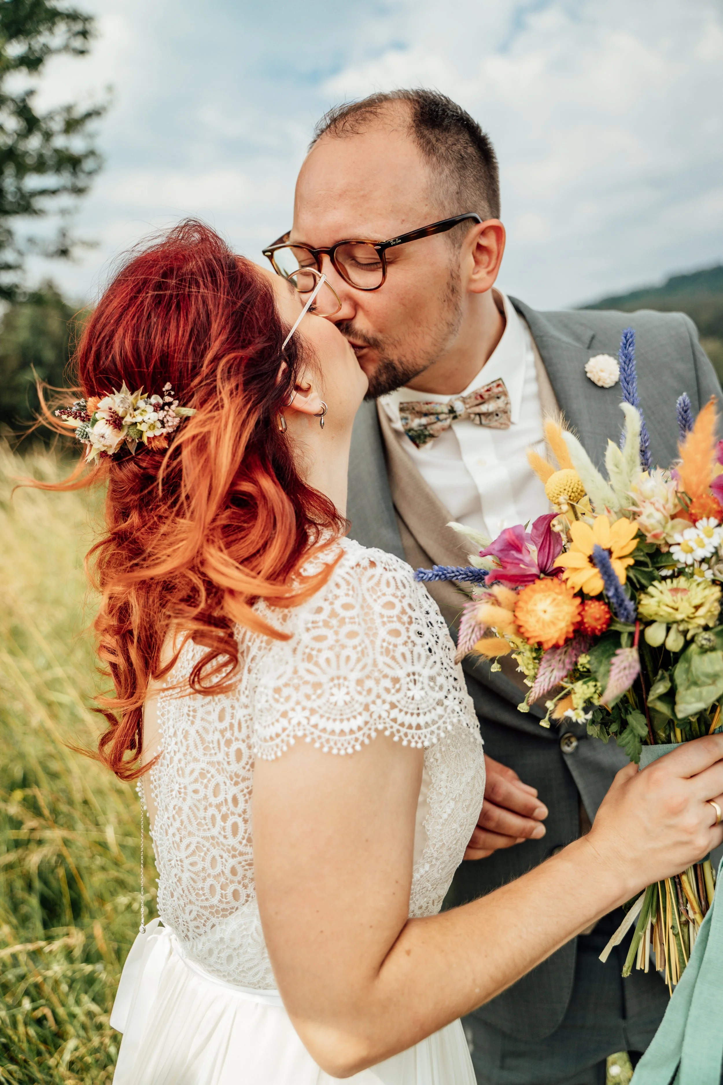 Ein Brautpaar küsst sich im Freien bei Sonnenlicht, die Braut trägt ein weißes Kleid mit Spitze und Blumendekor, der Bräutigam trägt einen grauen Anzug mit Fliege, das Brautpaar hält einen bunten Blumenstrauß.