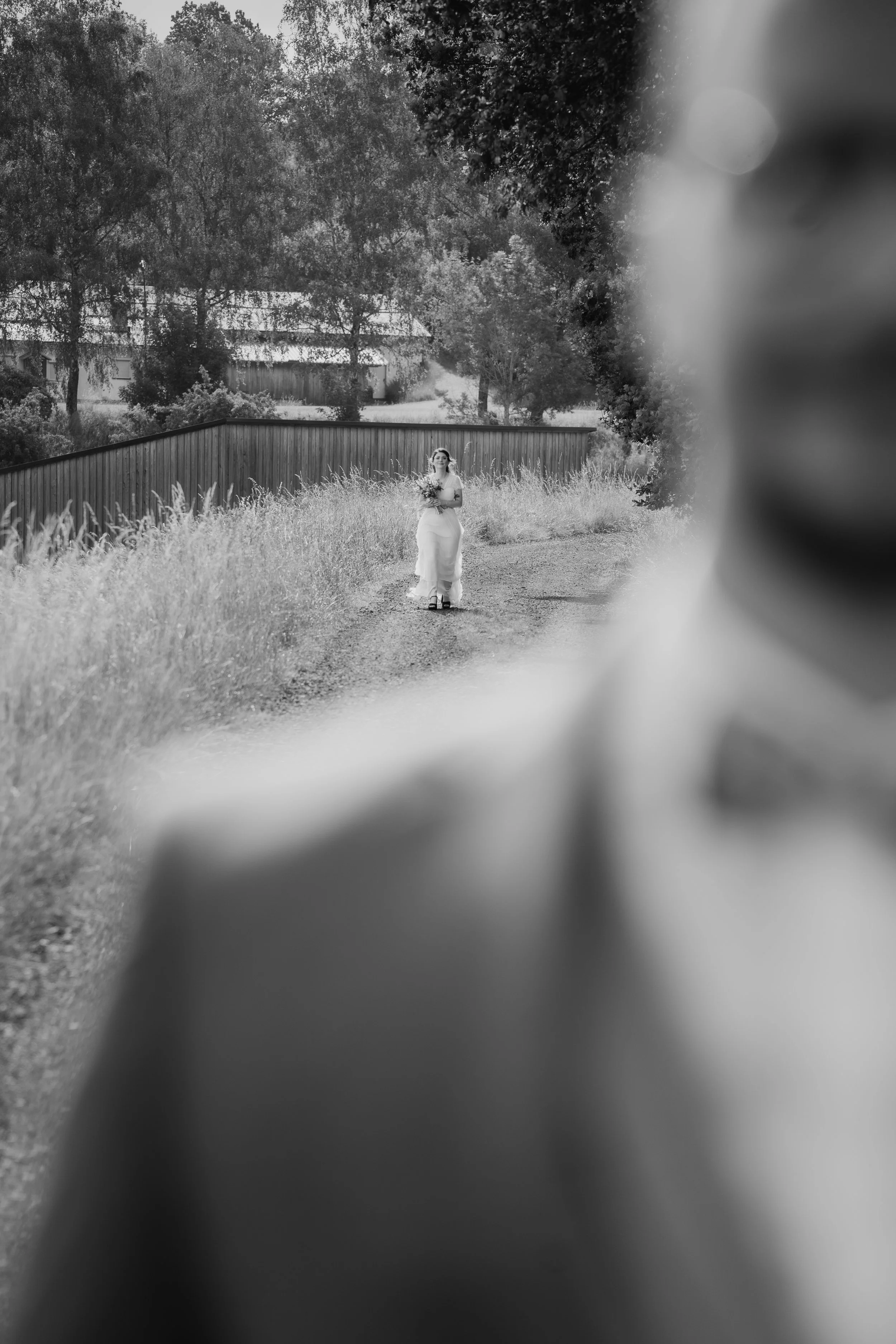 Eine Frau in einem weißen Kleid hält einen Blumenstrauß und läuft auf einem Weg im Freien, umgeben von Büschen und Bäumen, im Hintergrund ein Gebäude. Das Bild ist in Schwarz-Weiß.