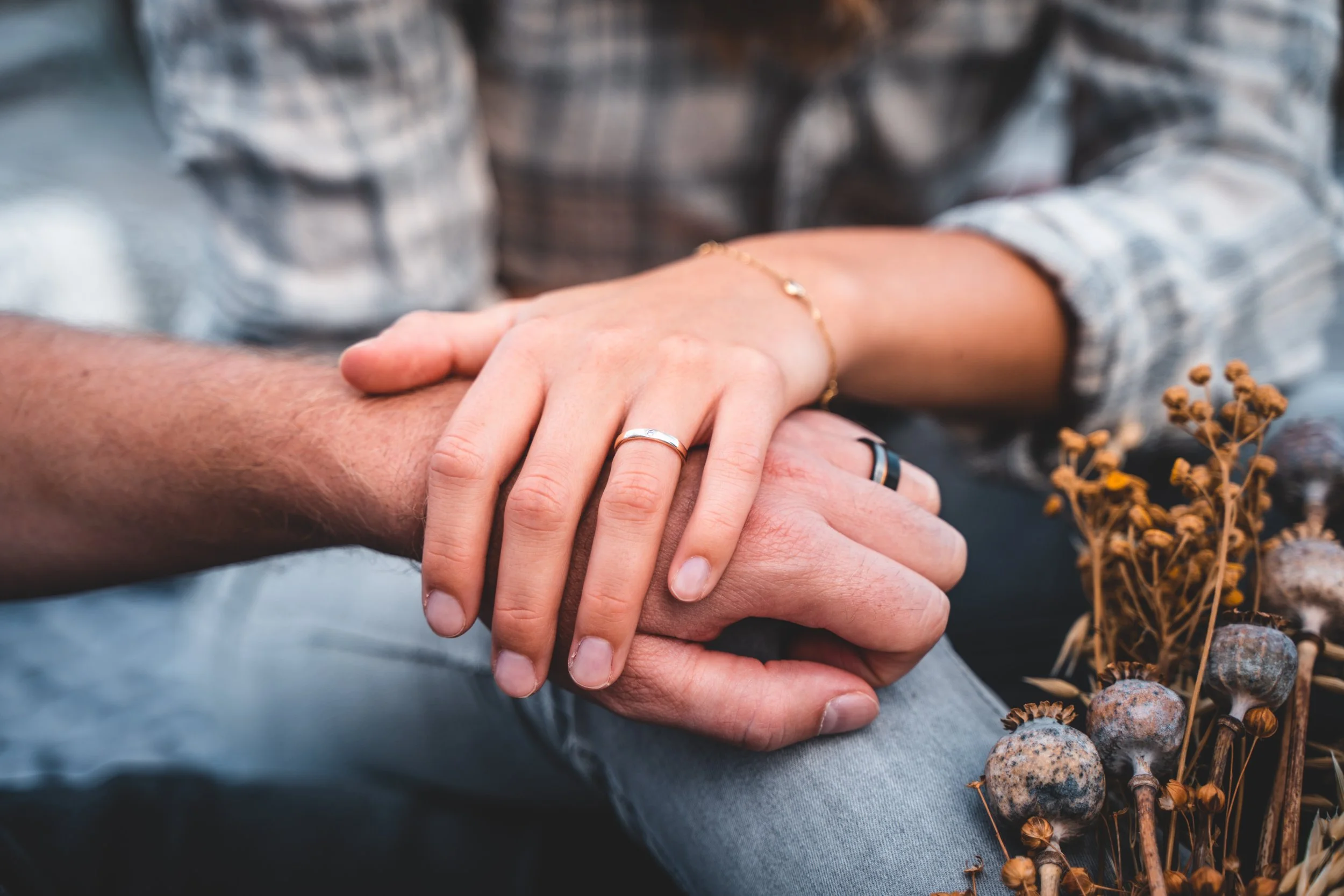Zwei Menschen halten sich bei einem Treffen die Hände, beide tragen Ringe und Armbänder, im Hintergrund befinden sich Trockenpflanzen und Steine.
