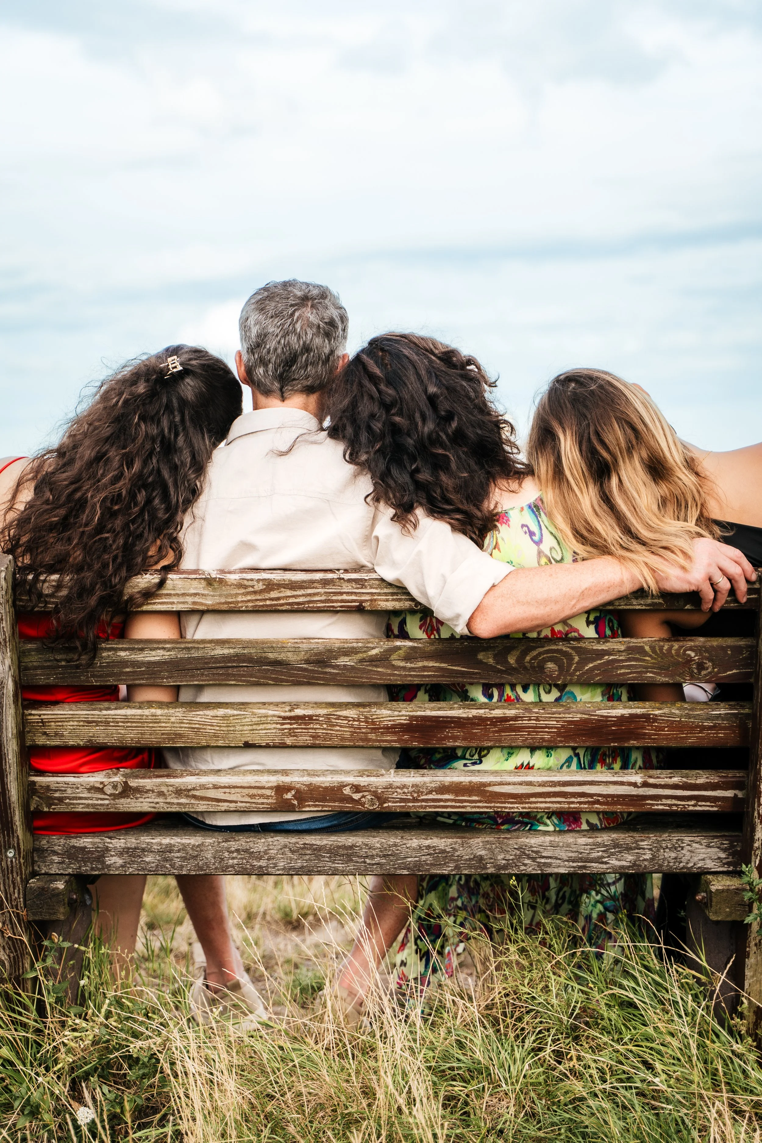 Vier Menschen sitzen auf einer Parkbank, mit dem Rücken zur Kamera, im Freien bei bewölktem Himmel. Es sind drei Frauen mit langen Haaren und ein Mann mit kurzen, grauen Haaren.