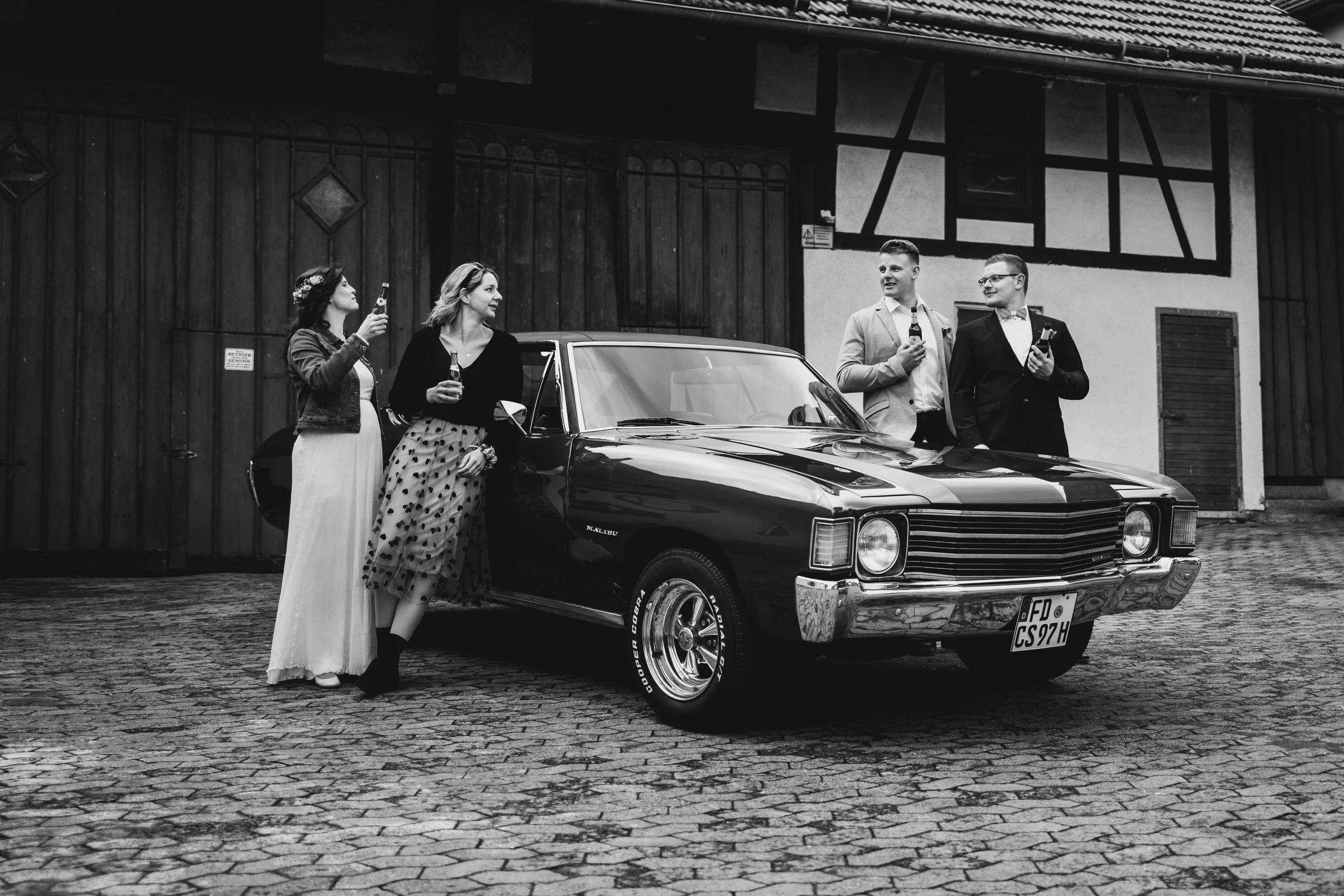 Vier Menschen, zwei Frauen und zwei Männer, stehen neben einem klassischen schwarzen Auto auf einem gepflasterten Platz vor einer alten Holztür. Alle halten Getränke und scheinen in einer entspannten Unterhaltung zu sein.