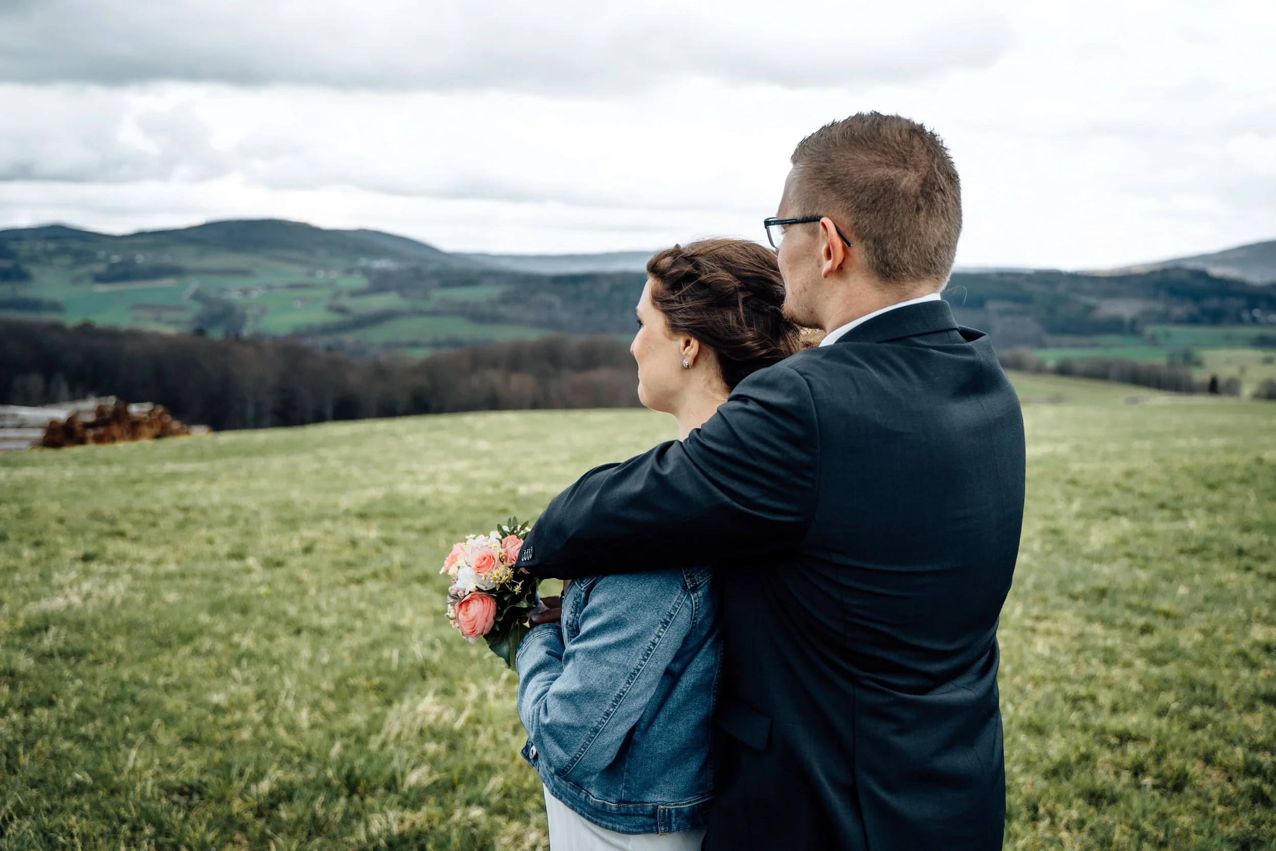 Ein Paar in formeller Kleidung sitzt im Freien auf grünem Gras, mit Bergen und einem bewölkten Himmel im Hintergrund, und hält sich bei einem regnerischen oder bewölkten Tag.