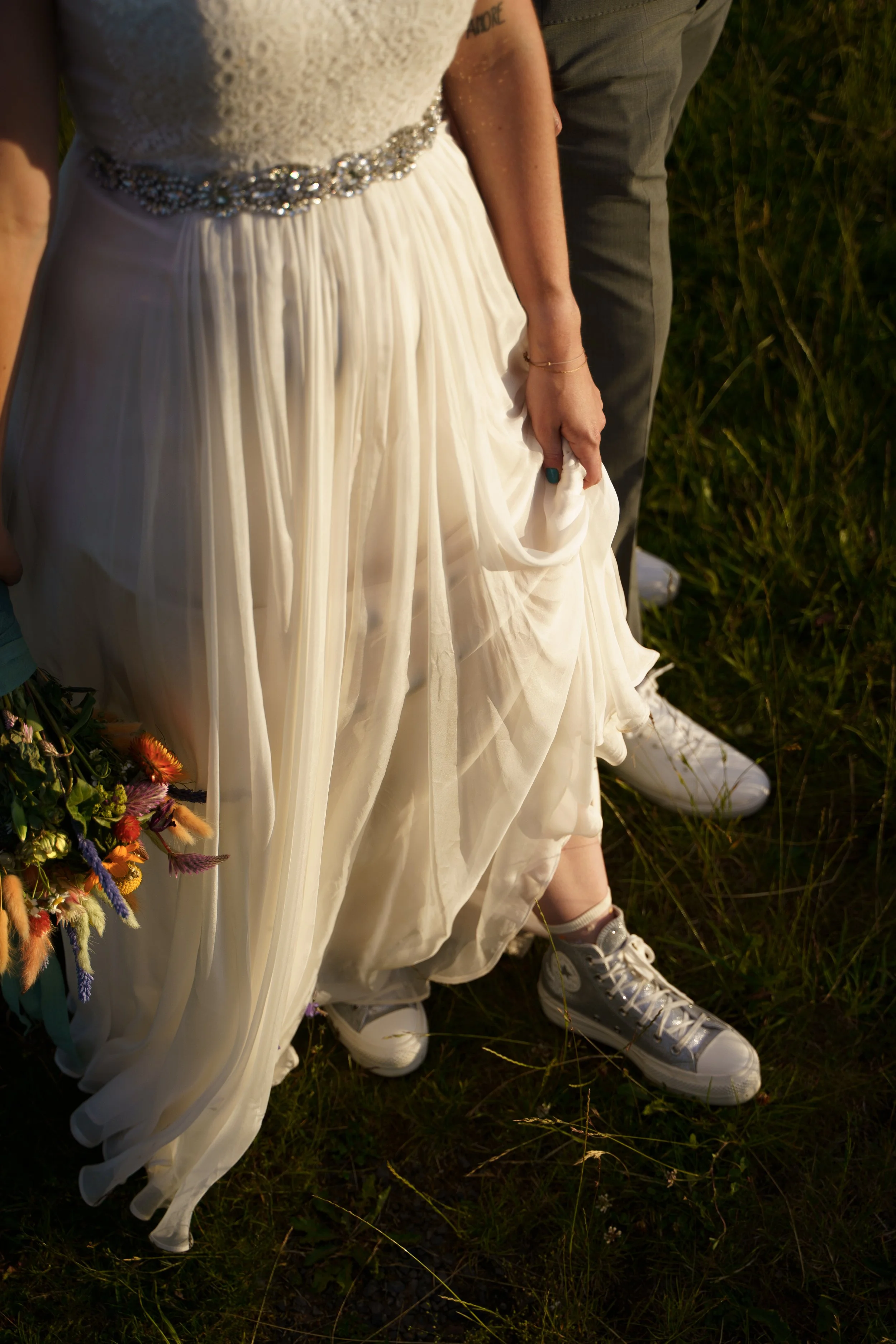 Nahaufnahme eines Brautpaares, der die Füße im Gras zeigt, mit weißen Sneakern und einem langen, weißen Brautkleid mit silberner Verzierung an der Taille.