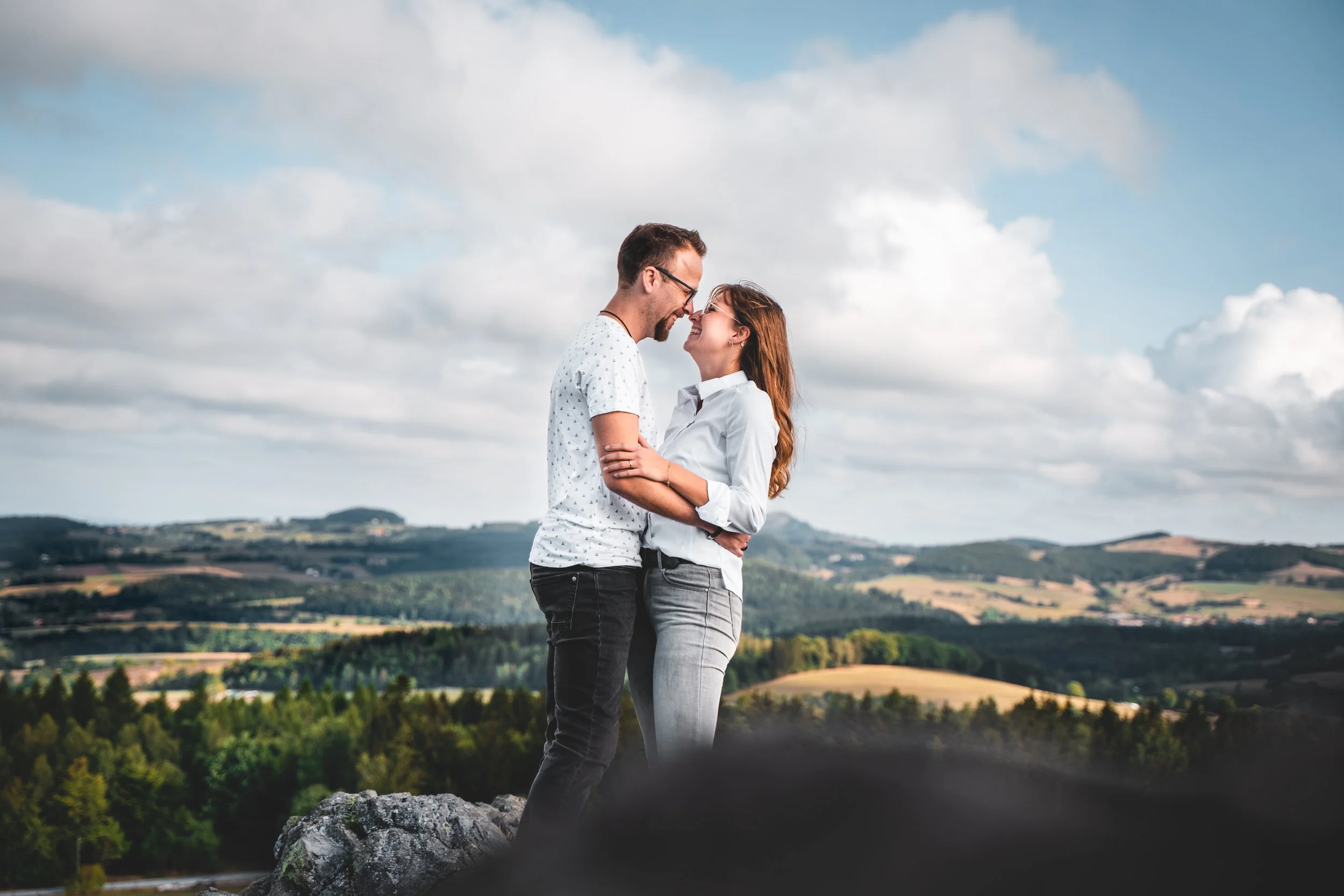 Ein Mann und eine Frau stehen sich lächelnd gegenüber, berühren sich an der Nasenspitze, vor einer ländlichen Landschaft mit Hügeln, Bäumen und einem bewölkten Himmel.