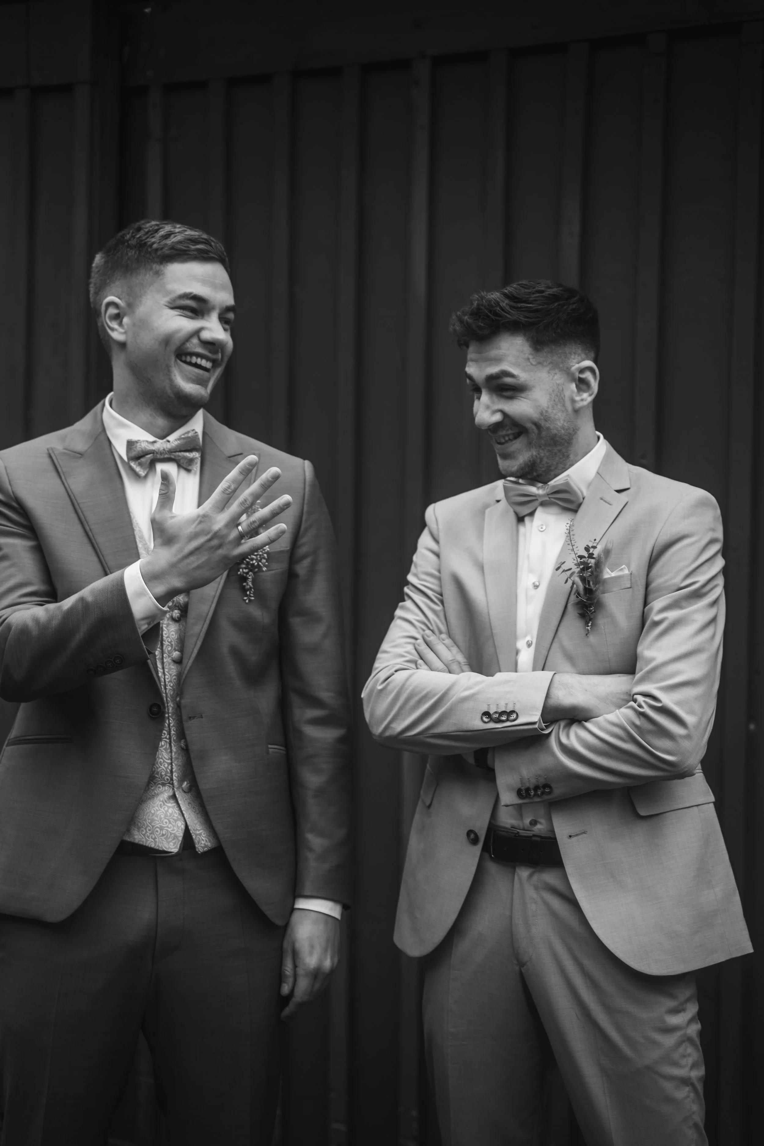 Zwei Männer in Anzügen lachen und unterhalten sich. Einer zeigt auf seine Hand, während der andere seine Arme verschränkt. Hintergrund aus dunklem Holz.