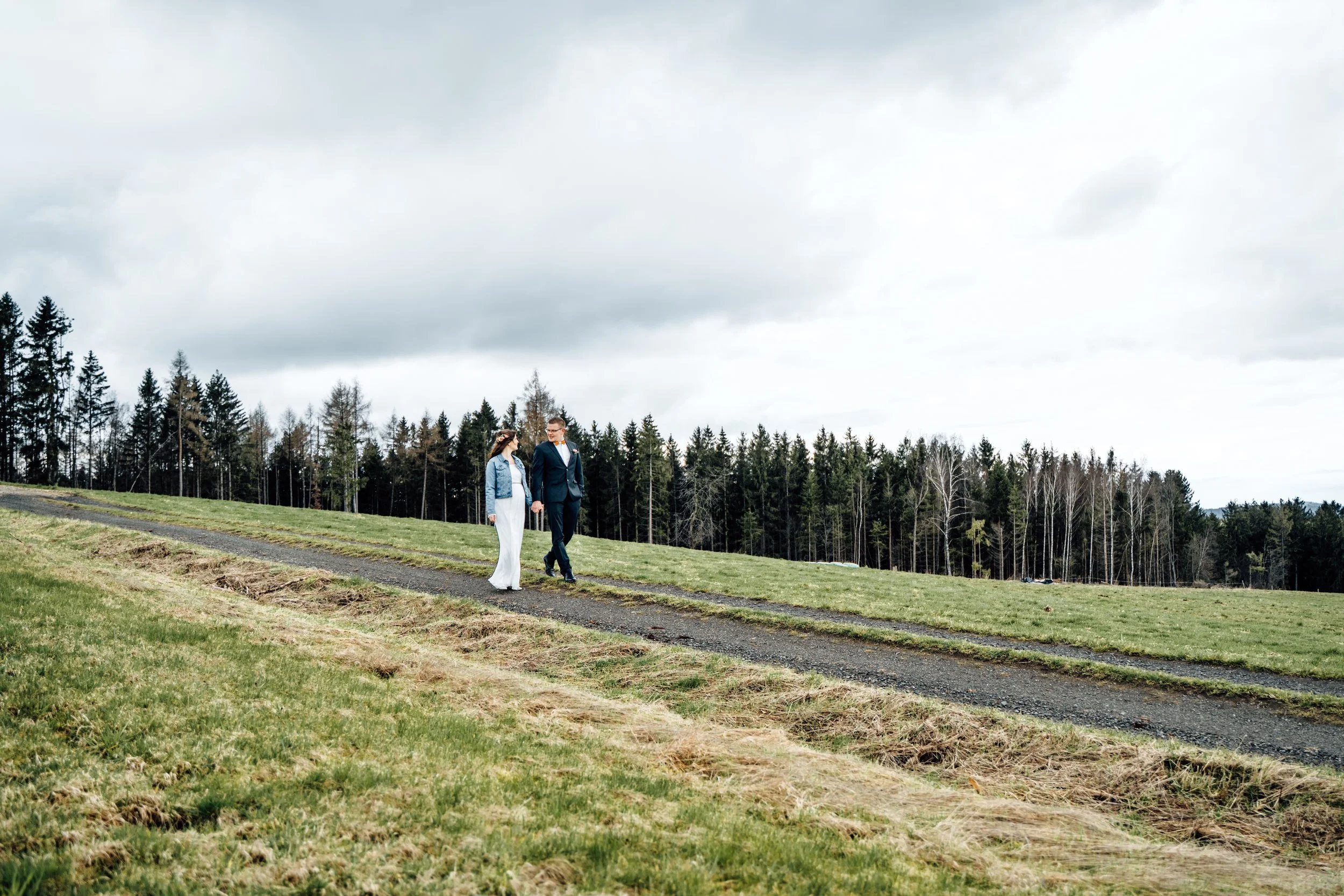 Ein Paar, wahrscheinlich Braut und Bräutigam, geht Hand in Hand auf einem Feld mit Gräsern, umgeben von einem Nadelwald, bei bewölktem Himmel.