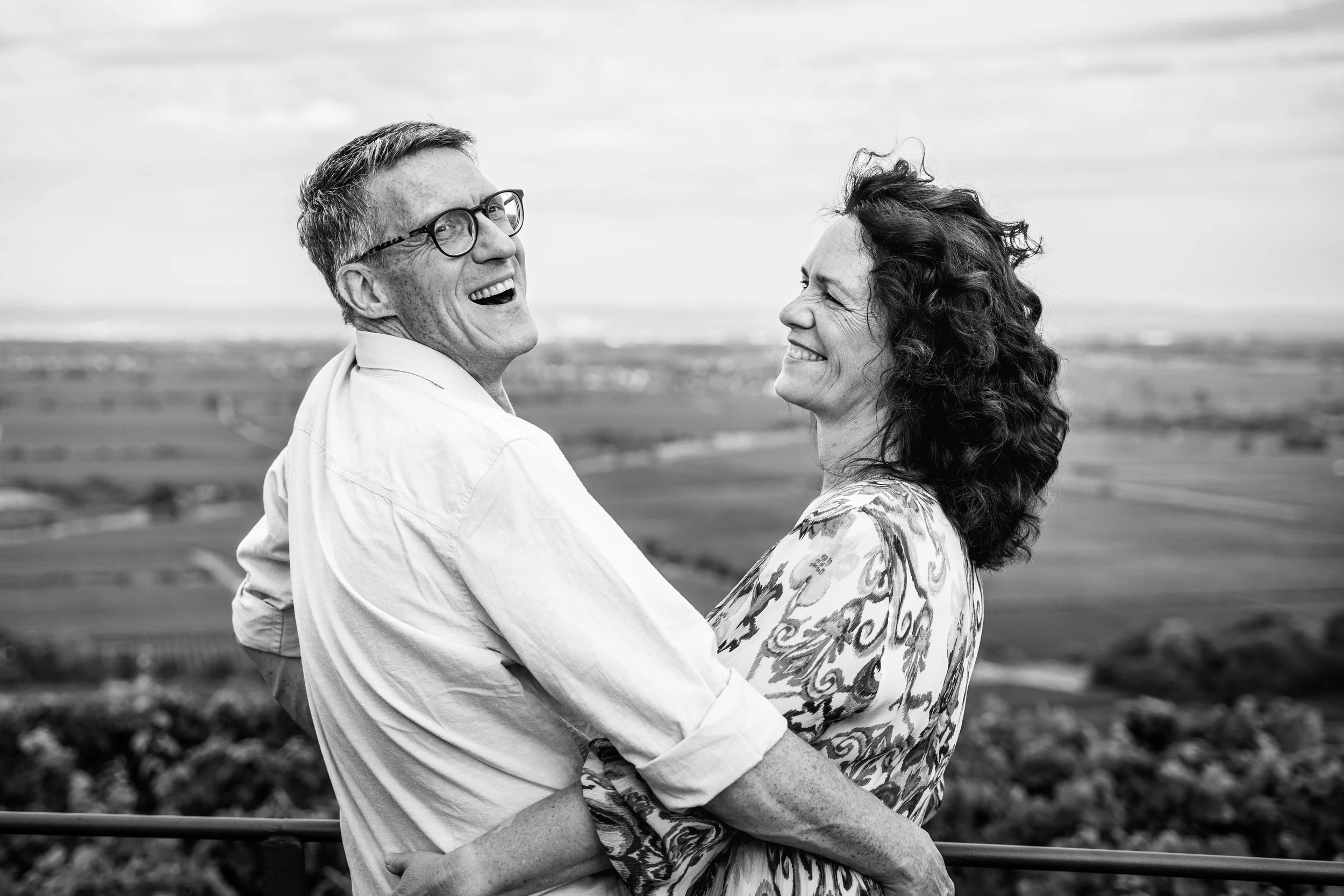 Ein lachendes Paar, ein Mann mit Brille und eine Frau, die sich umarmen, im Freien mit Landschaft im Hintergrund.