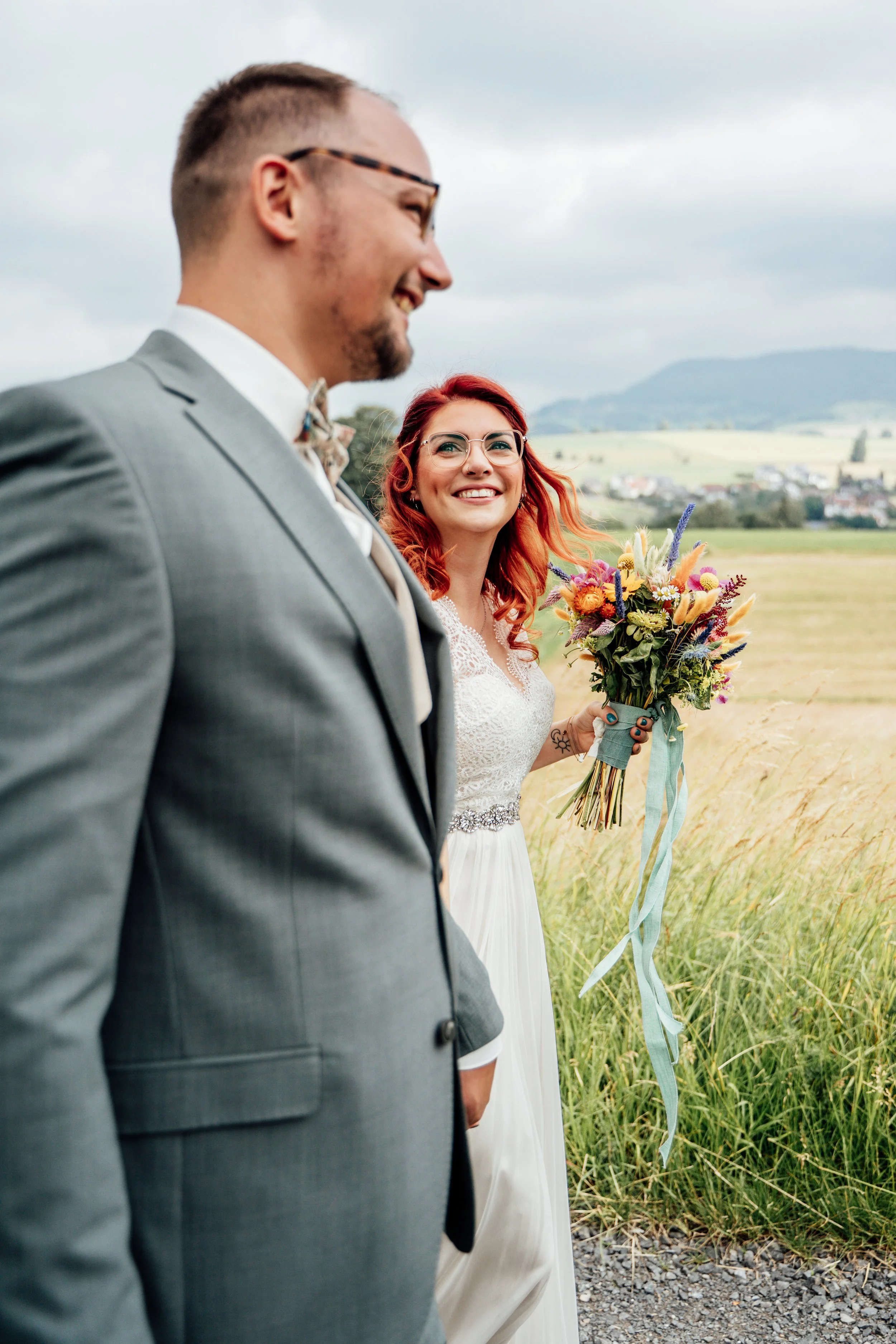 Ein Brautpaar steht in einem Feld im Freien, die Frau hält einen bunten Blumenstrauß, beide sind in festlicher Kleidung, im Hintergrund eine ländliche Landschaft mit Bergen und Wolken am Himmel.
