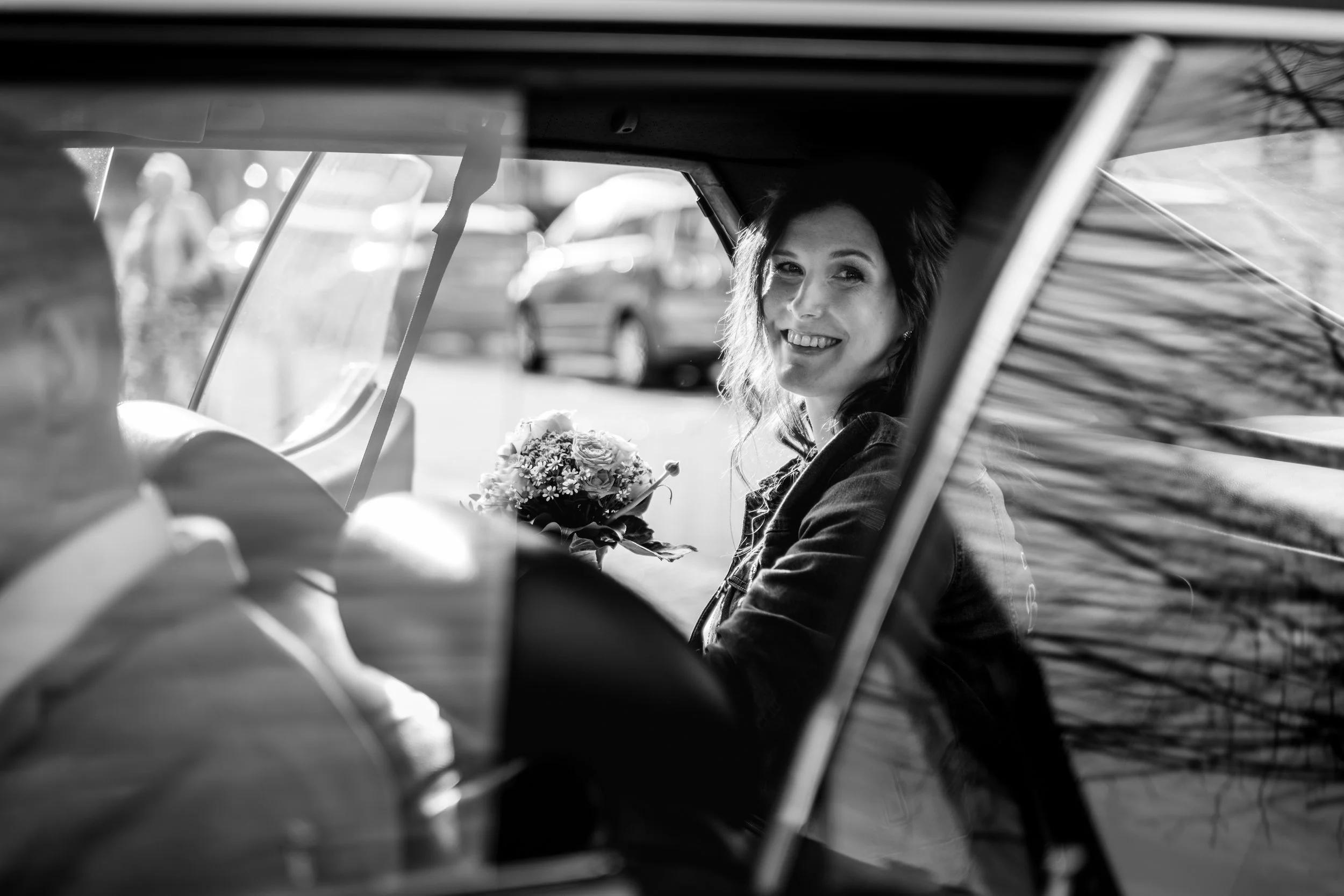 Eine lachende Frau mit dunklen, gewellten Haaren sitzt in einem Auto, hält einen Blumenstrauß in der Hand und schaut aus dem Fenster. Das Bild ist in Schwarz-Weiß.