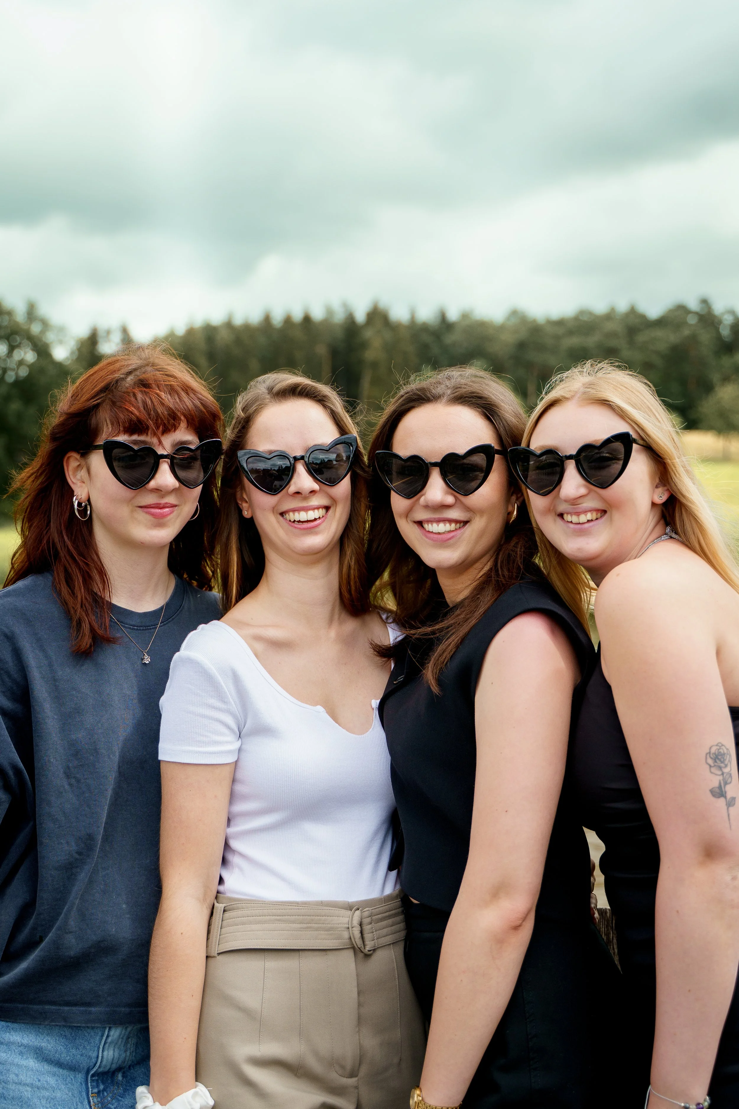 Vier Frauen mit Sonnenbrillen, die sich vor einer grünen Wiese und einem bewölkten Himmel befinden.