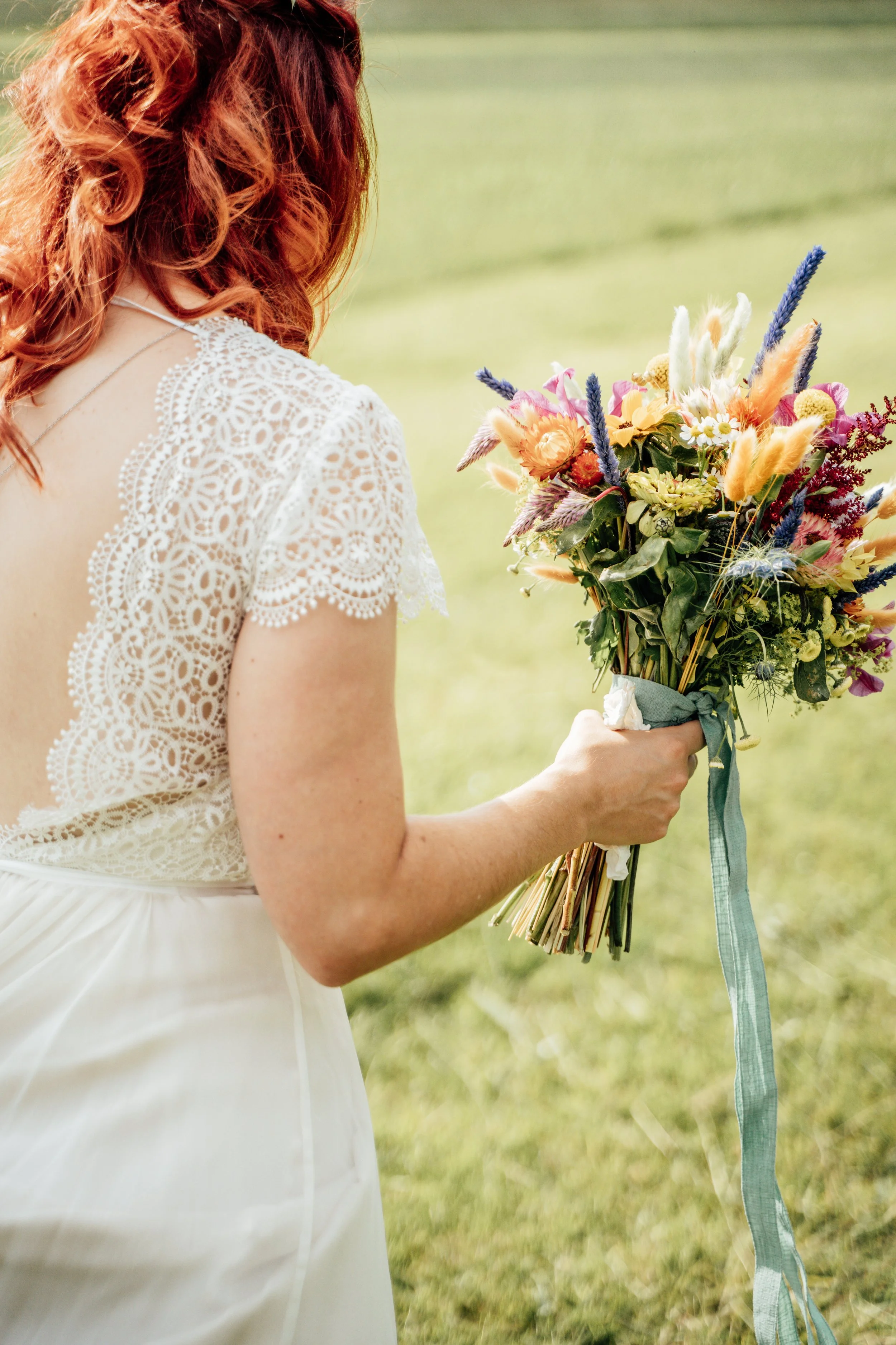 Eine junge Frau in einem weißen Kleid mit Spitzendetail hält einen bunten Blumenstrauß in der Hand, steht im Freien auf einer Wiese.