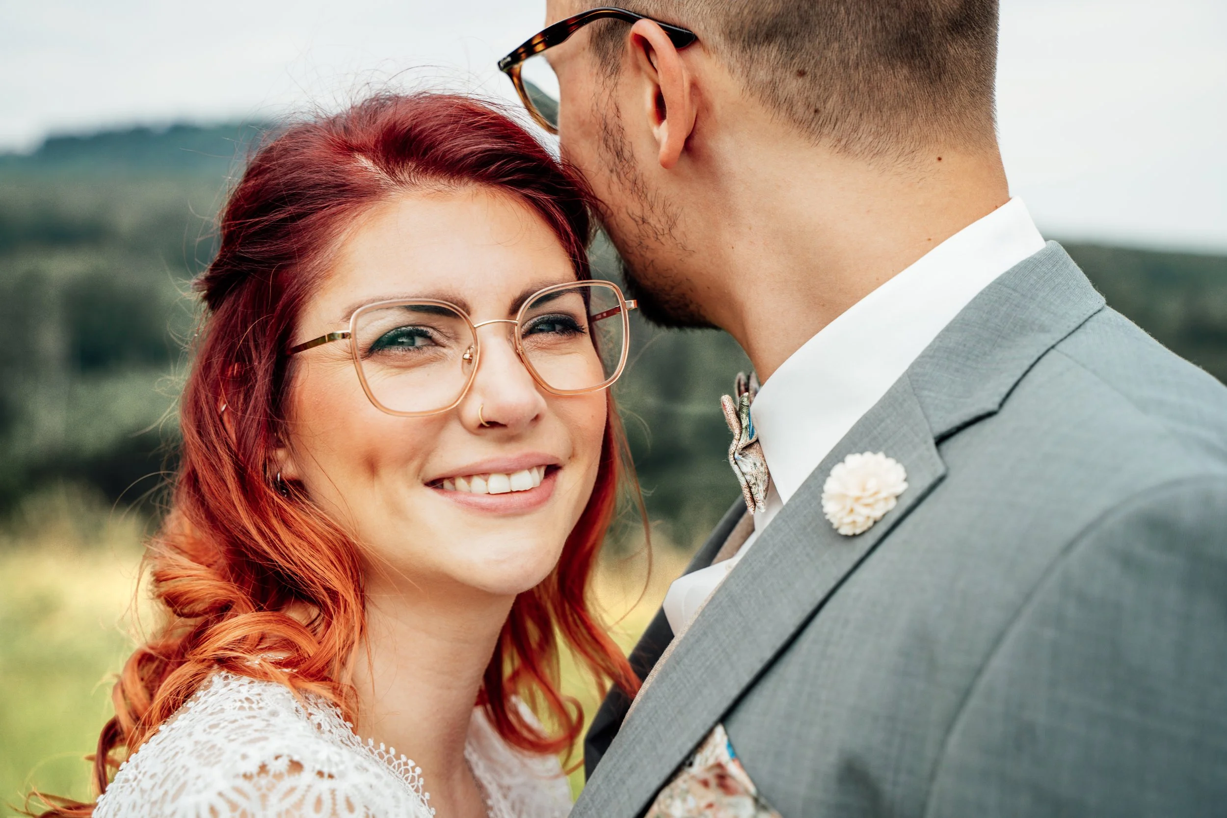 Ein Paar im Hochzeitsoutfit, die sich liebevoll berühren und küssen, vor einer natürlichen, grünen Landschaft.