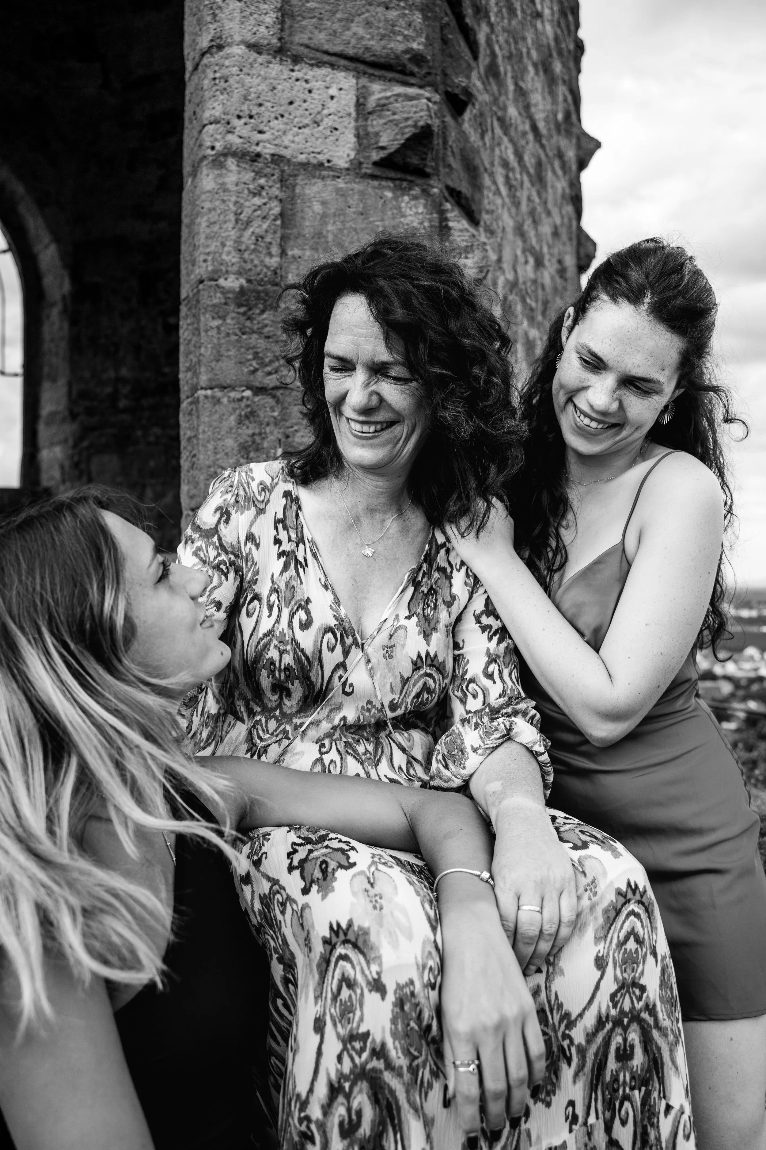 Drei Frauen lachen und umarmen sich vor einer alten Steinmauer, im Schwarz-Weiß-Format.