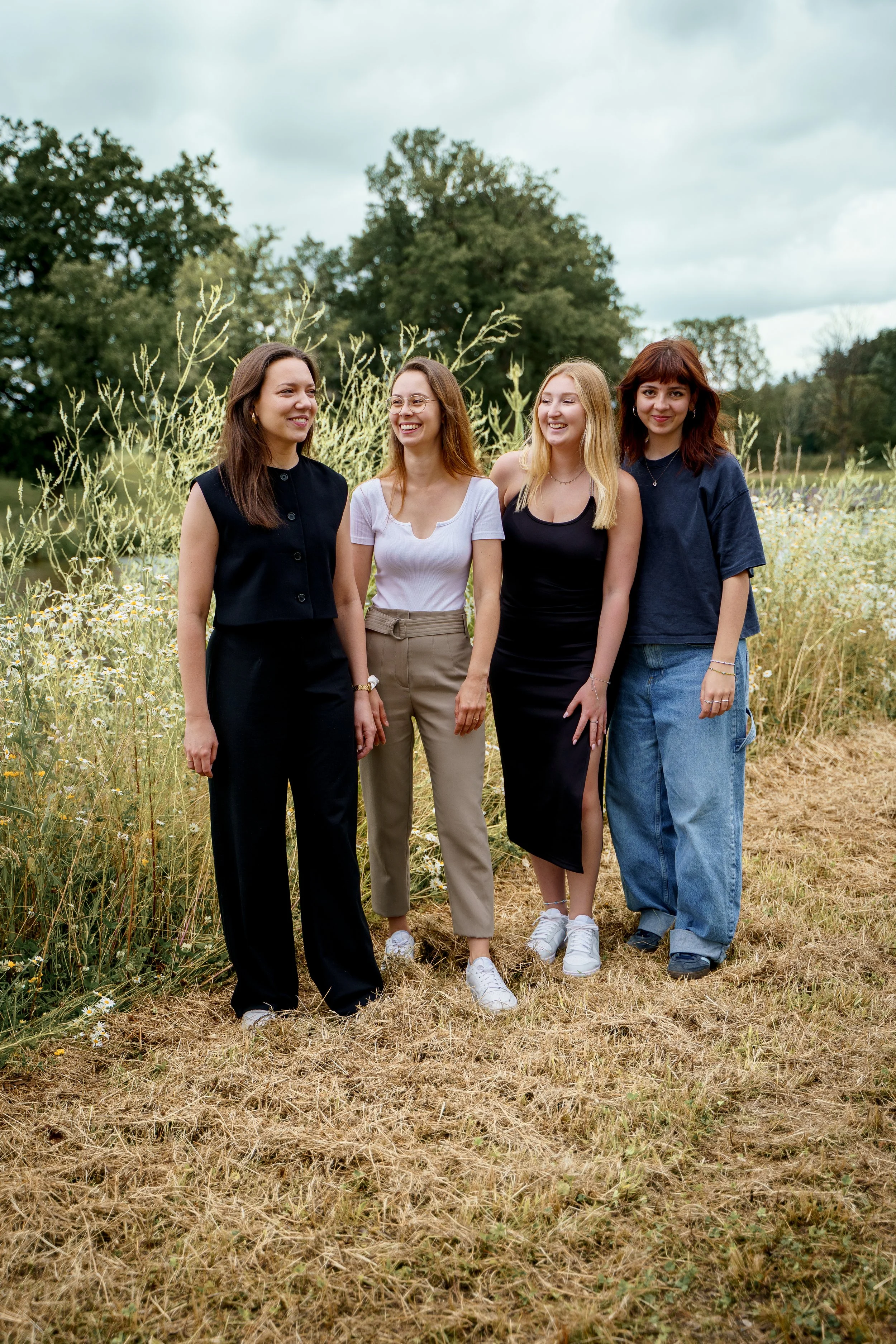 Vier junge Frauen stehen in einem Feld mit Gräsern und Blumen, lachen und genießen das sonnige Wetter, im Hintergrund sind Bäume und ein bewölkter Himmel sichtbar.