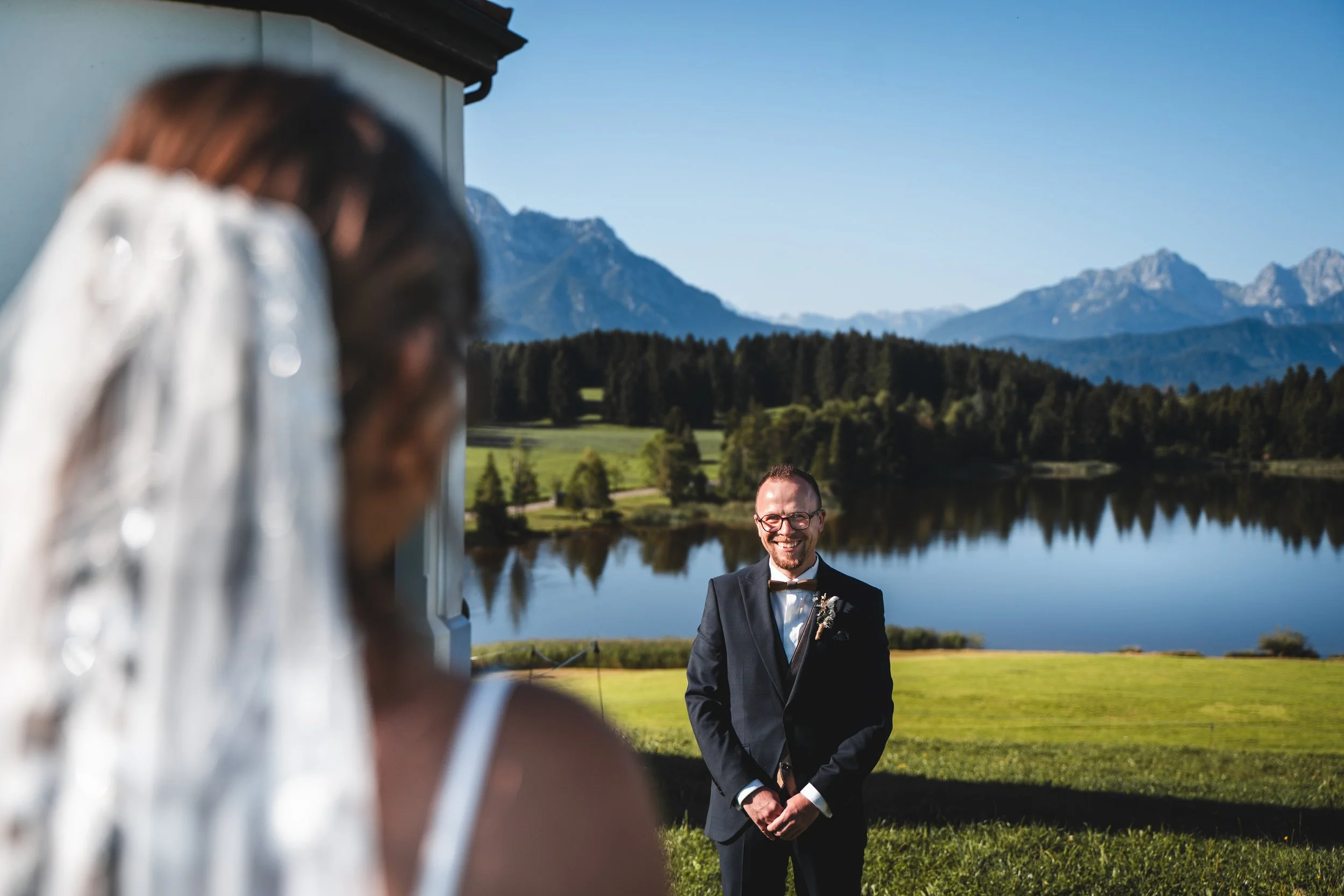 Ein Mann in formeller Kleidung steht vor einem See mit Bergen im Hintergrund, während eine Person im Vordergrund unscharf fotografiert wird.
