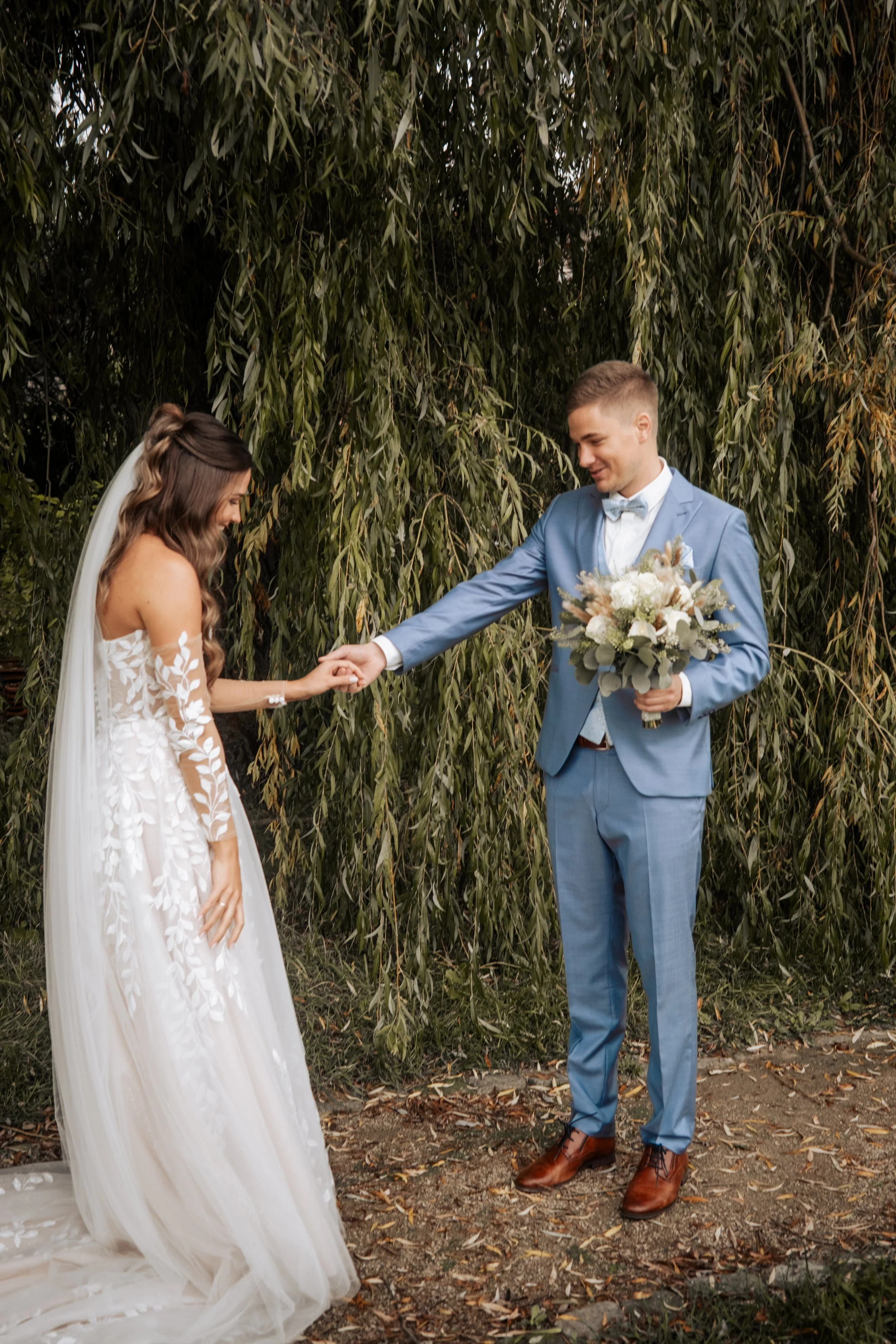 Ein Brautpaar bei einer Hochzeit im Freien, der Bräutigam reicht der Braut die Hand, im Hintergrund Bäume und Laub auf dem Boden.