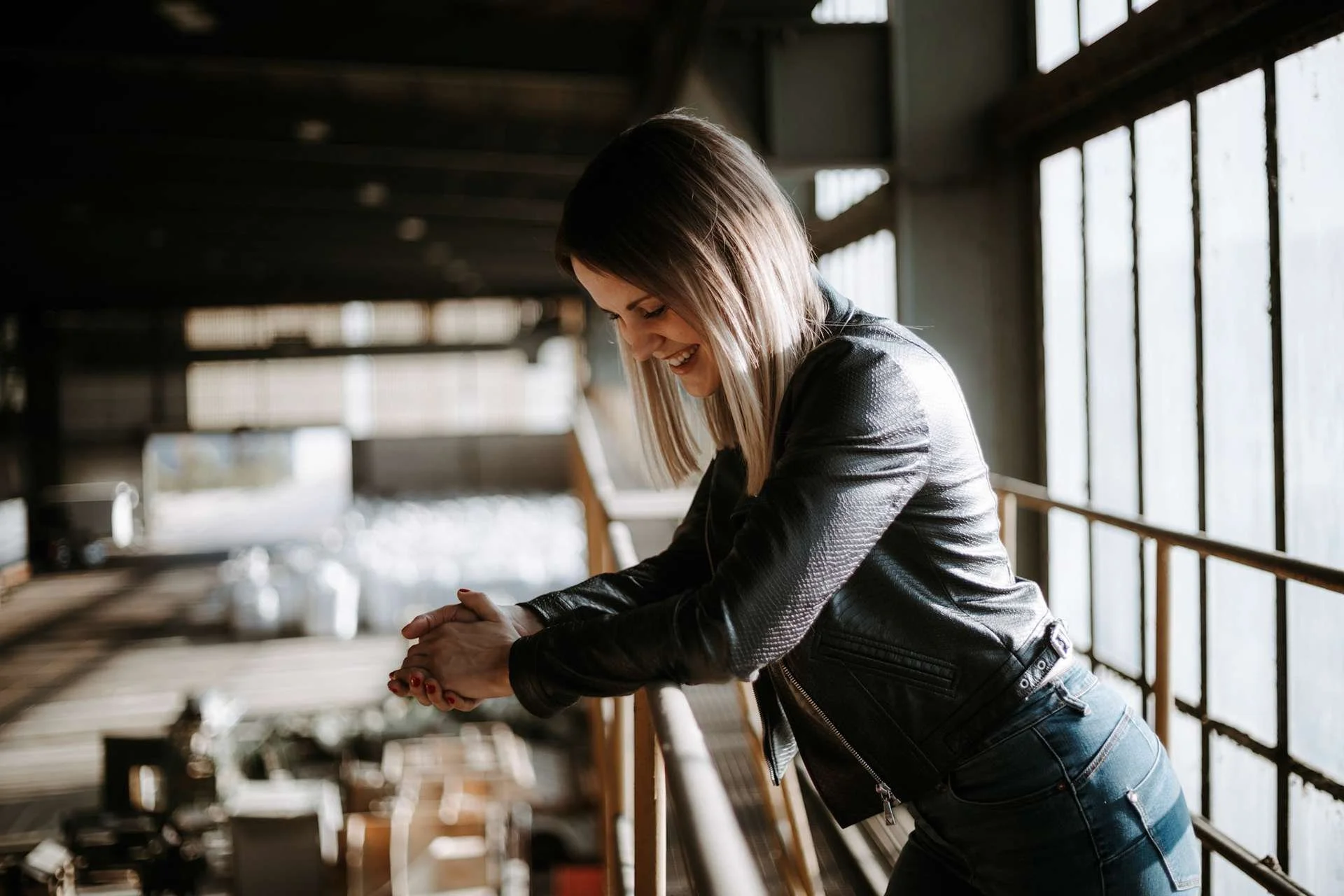 Junge Frau mit dunklem Haar in Lederjacke, lächelnd, in einem industrialartigen Raum mit großen Fenstern, lehnt an einem Geländer.