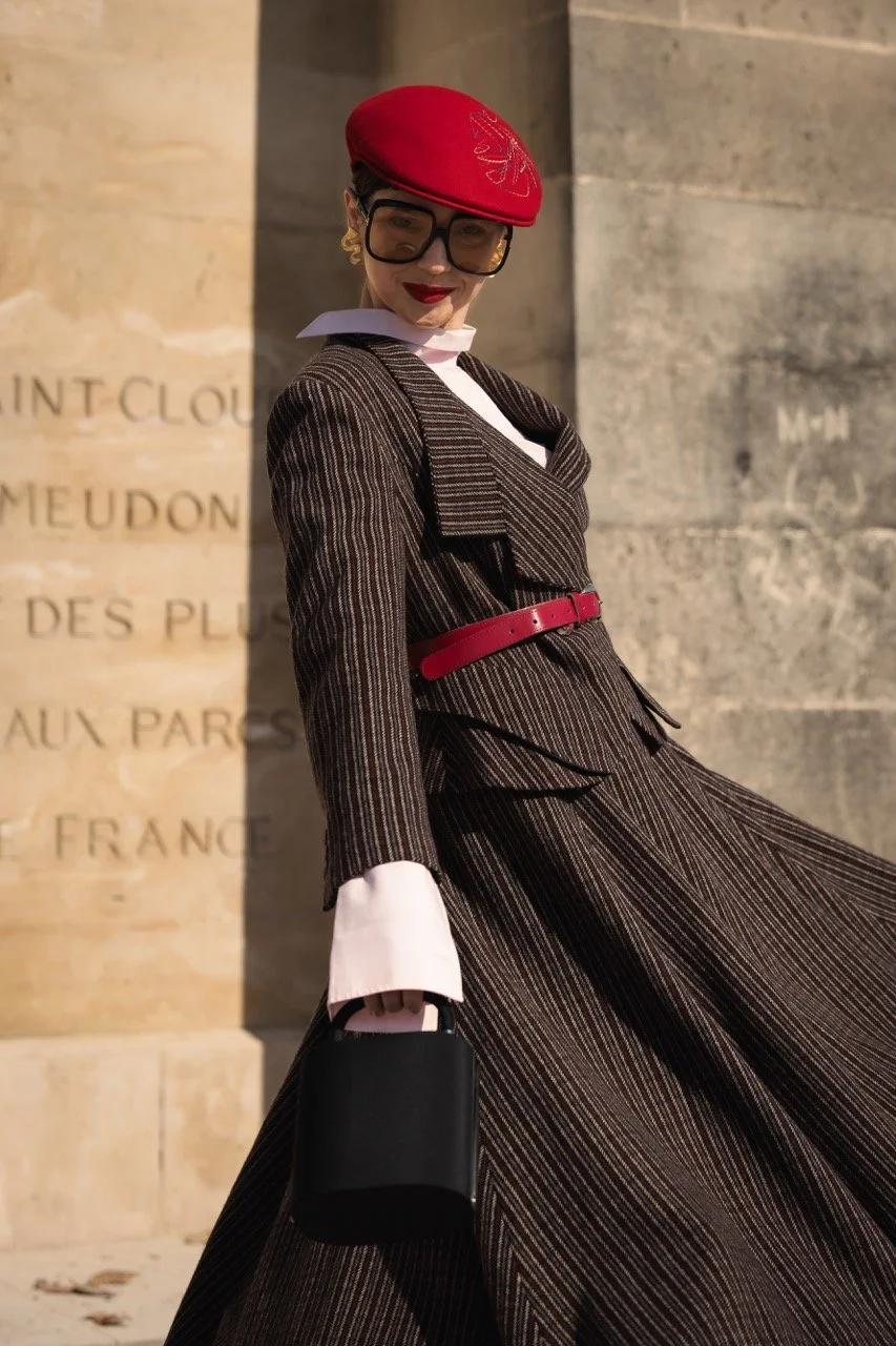 Frau in eleganter Karojacke, mit rotem Hut und schwarzer Tasche, vor einer Steinfassade.