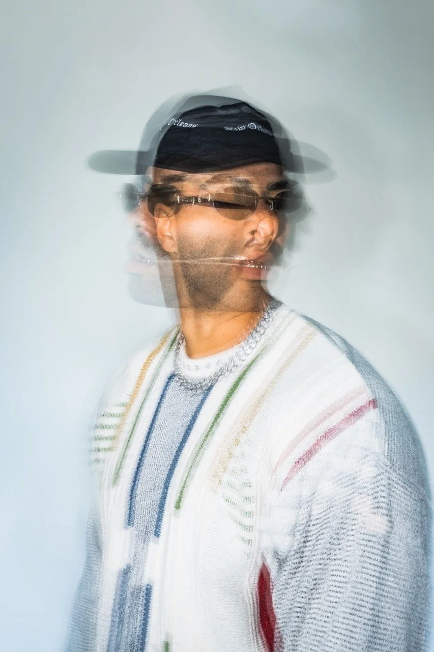 Ein Mann mit dunklem Baseballcap, Sonnenbrille und buntem gestricktem Pullover, der sich bewegt und daher unscharf und mehrfach im Bild dargestellt ist.