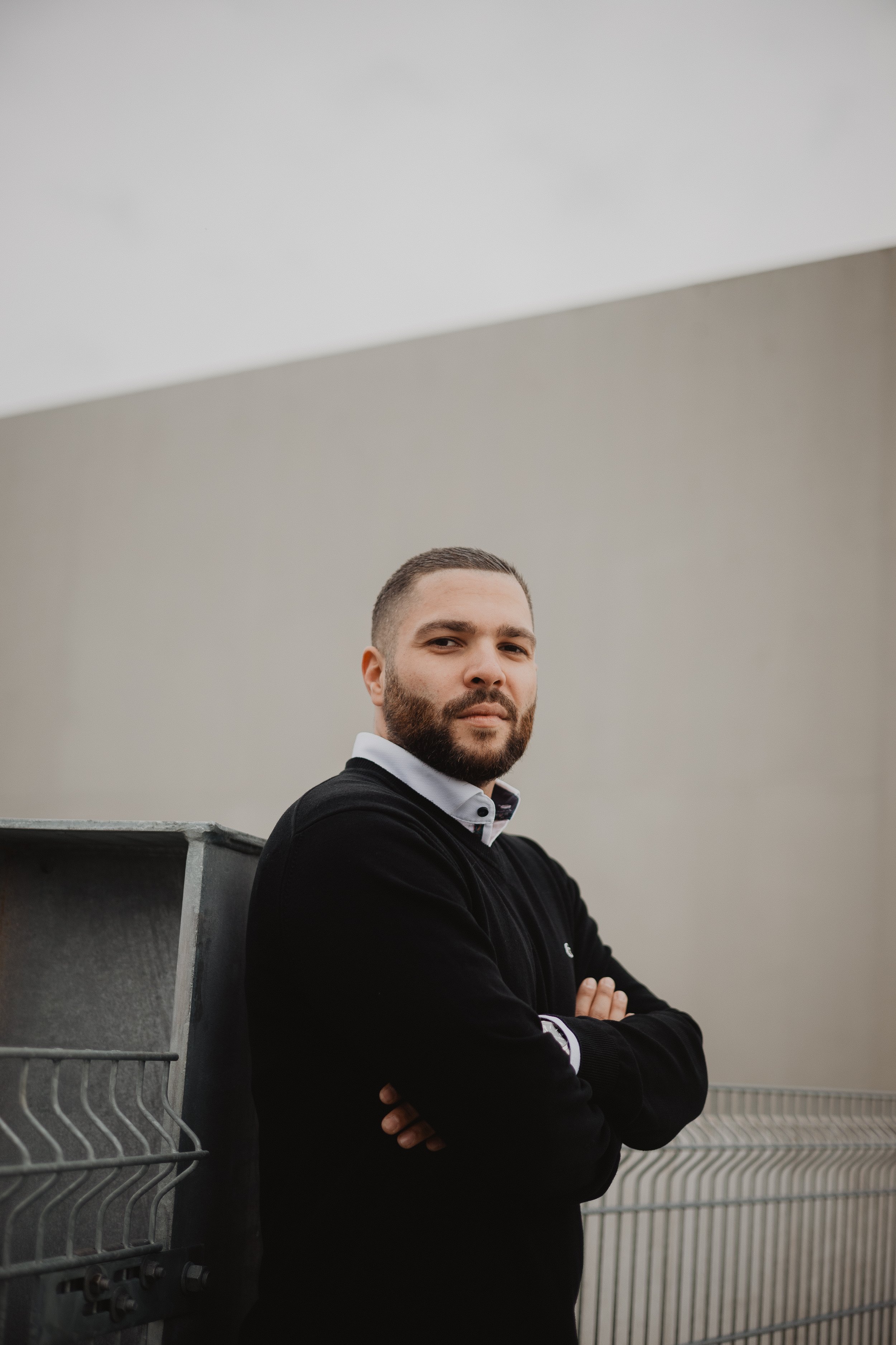 Ein Mann mit dunklem Pullover und weißen Hemd steht mit verschränkten Armen vor einer grauen Wand und einem Metallcontainer.