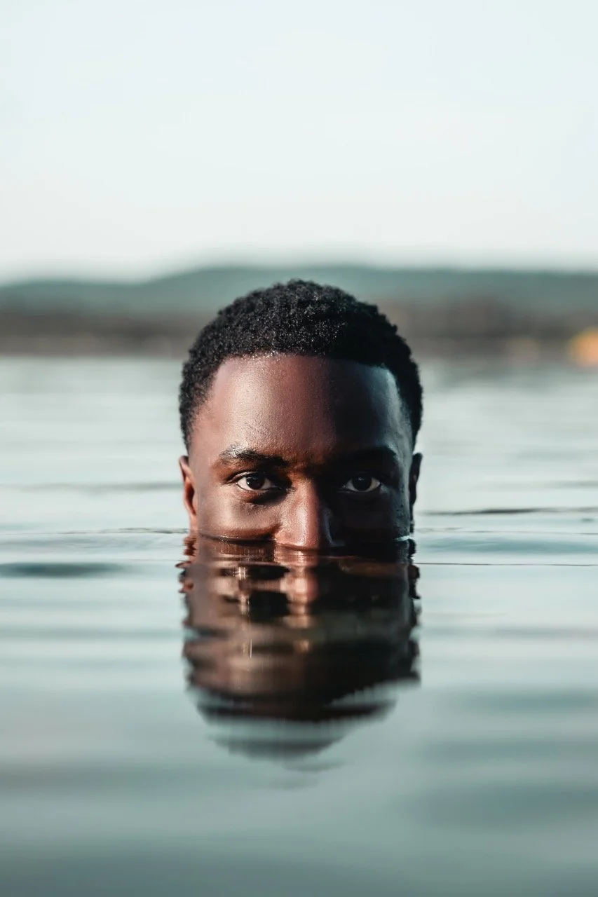 Man taucht im Wasser mit nur Kopf sichtbar, Wasser spiegelt seine Augen und Stirn, Hintergrund unscharf, sunsetlicht.