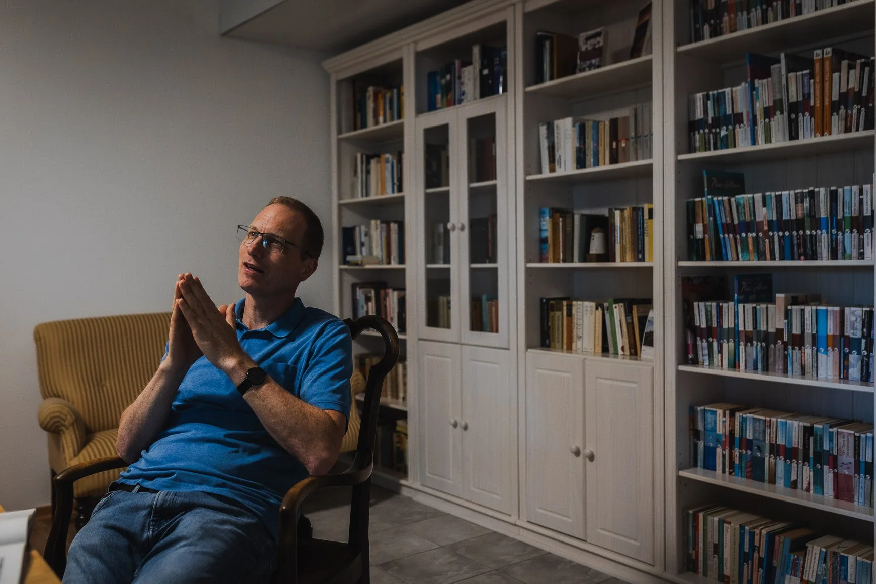 Ein Mann sitzt in einem blauen T-Shirt in einem Wohnzimmer, mit einem Bücherregal im Hintergrund.