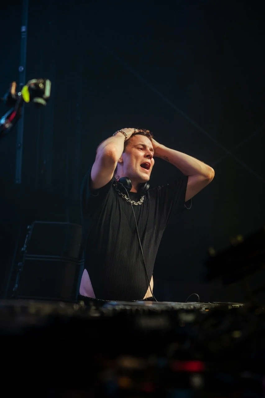 Ein junger Mann auf einem DJ-Pult, der erschöpft oder emotional ist, während er die Hände auf dem Kopf hat, bei einer Musikveranstaltung.