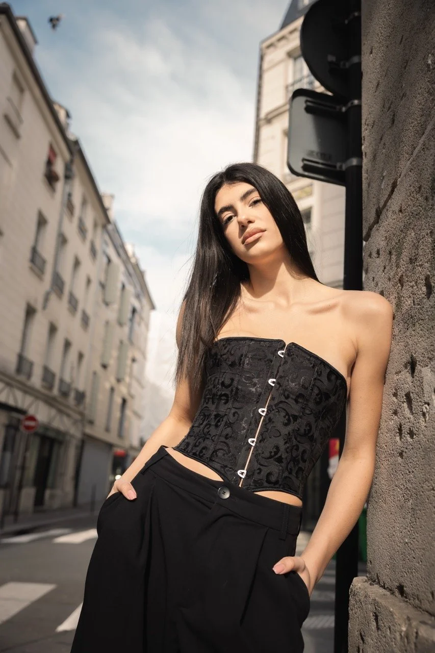 Junge Frau in schwarzem Korsett und schwarzen Hosen steht an einer Stadtstraße, lehnend an eine Steinmauer, bei Tageslicht