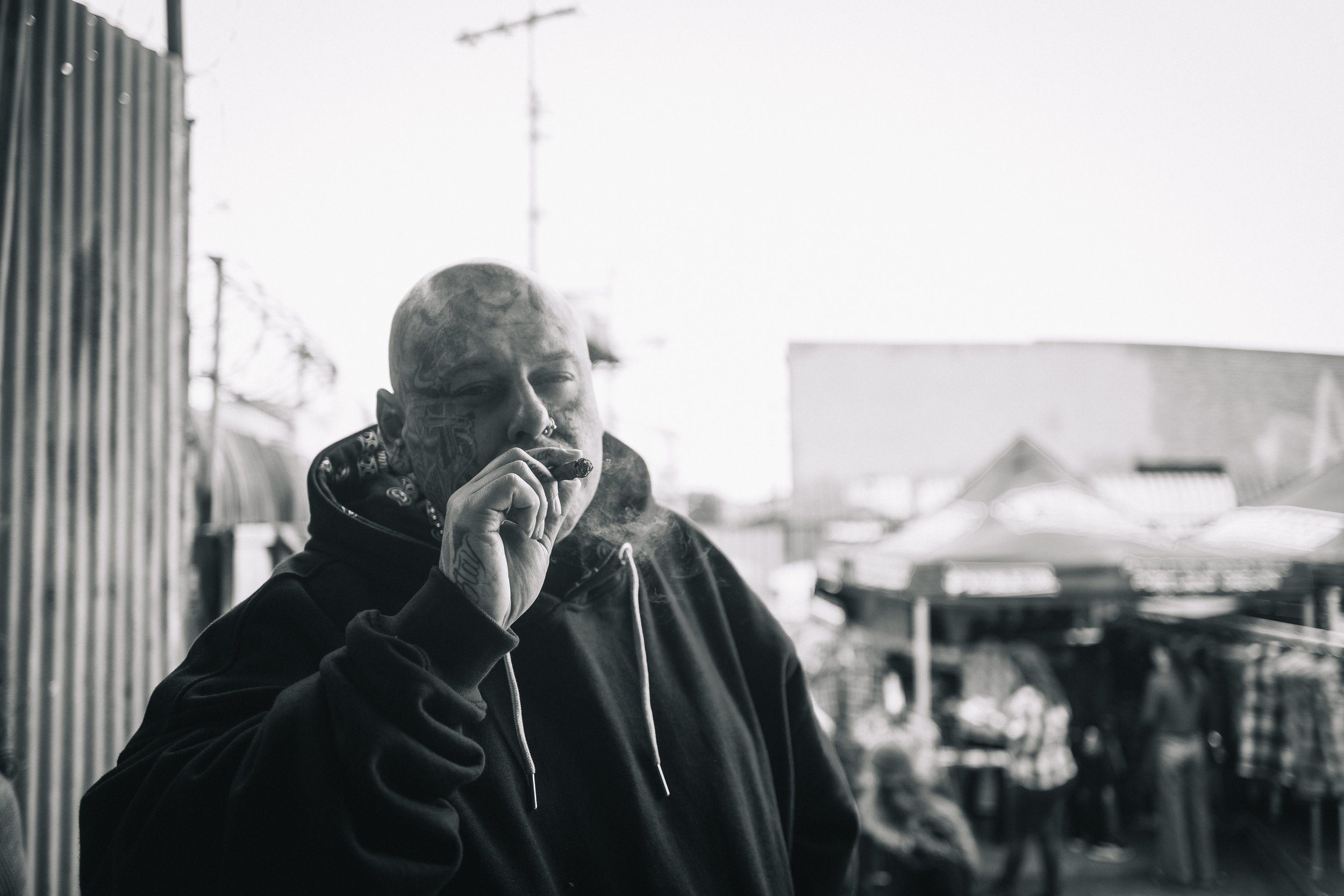 Ein tattooüberzogener Mann trägt einen schwarzen Kapuzenpullover und raucht eine Zigarre auf einem Balkon