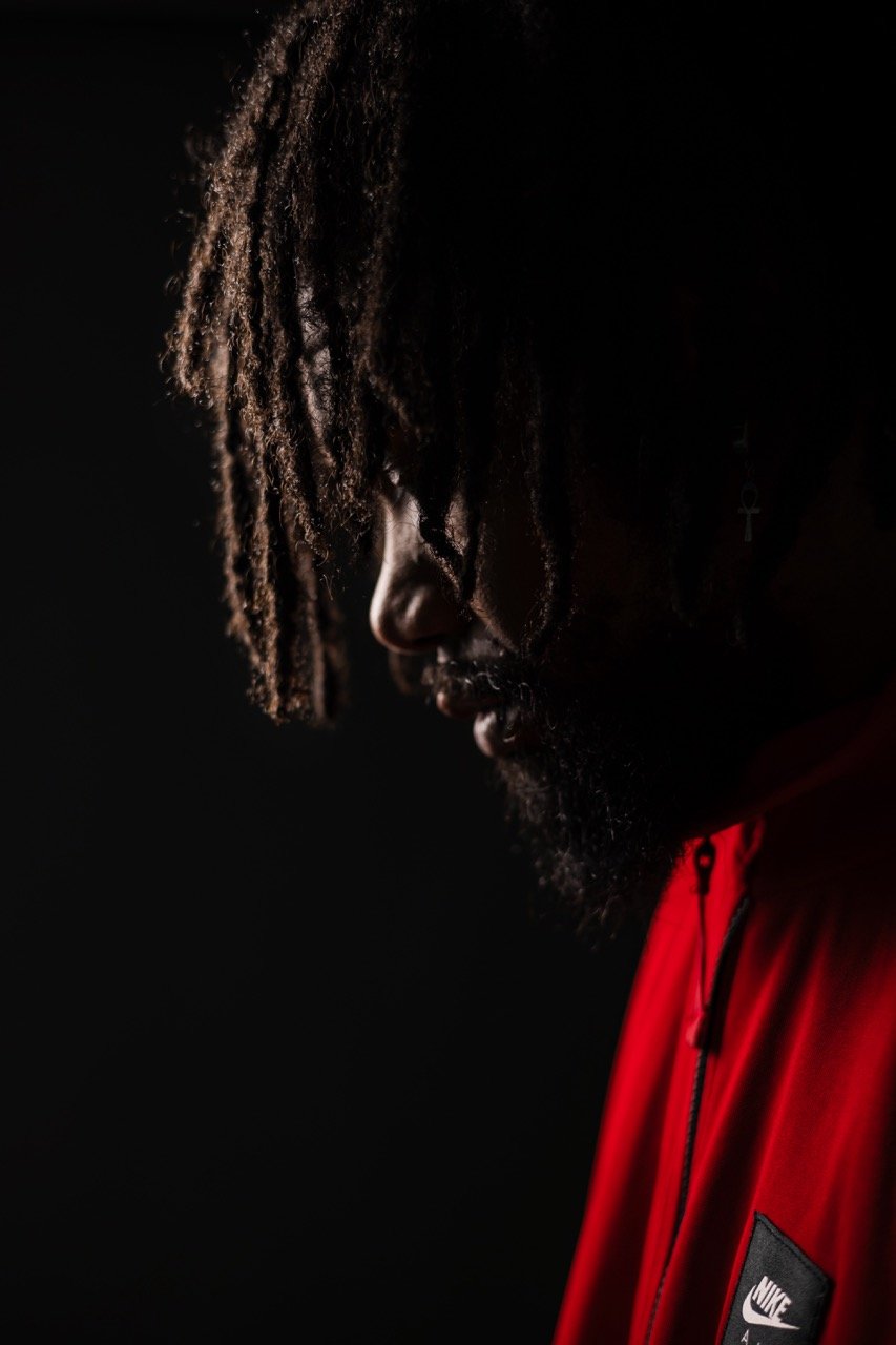 Nahaufnahme eines Mannes mit dreadlocks, dunklem Bart und rotem Nike-Jacket, sorgfältig beleuchtet vor dunklem Hintergrund.