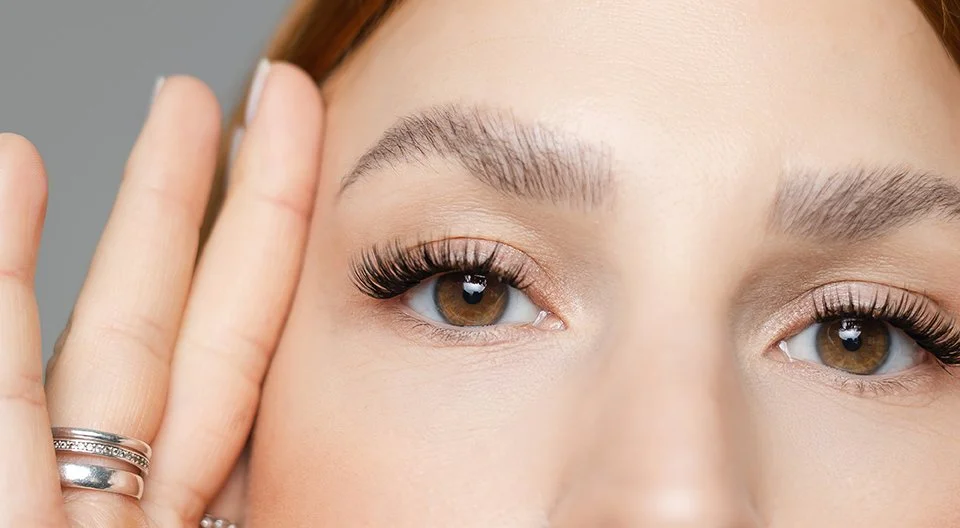 Nahaufnahme eines Frauengesichts mit braunen Augen, geschminkten Wimpern und gepflegten Brauen, die ihre Hand mit mehreren Ringen zeigt.