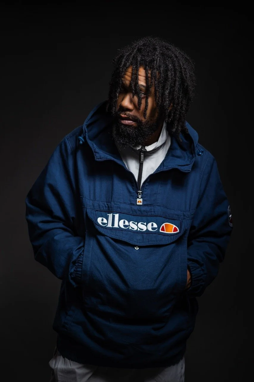 Junge männliche Person mit Dreadlocks in dunklem Studio, trägt eine blaue Ellesse-Jacke.