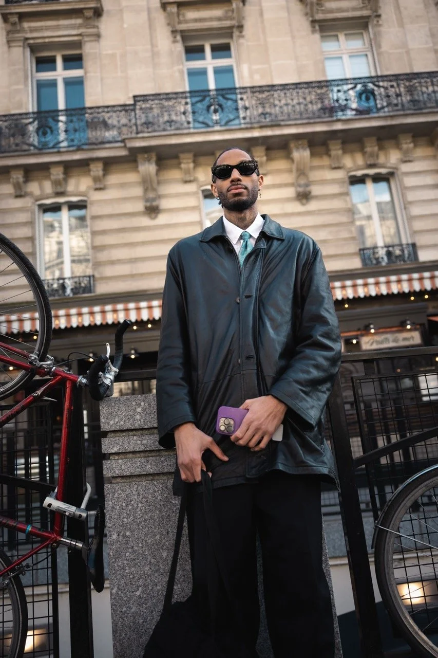 Ein Mann steht vor einer Paris-typischen Gebäudefassade mit Balkon, trägt eine schwarze Lederjacke, dunkle Sonnenbrille und hält ein Smartphone in der Hand.