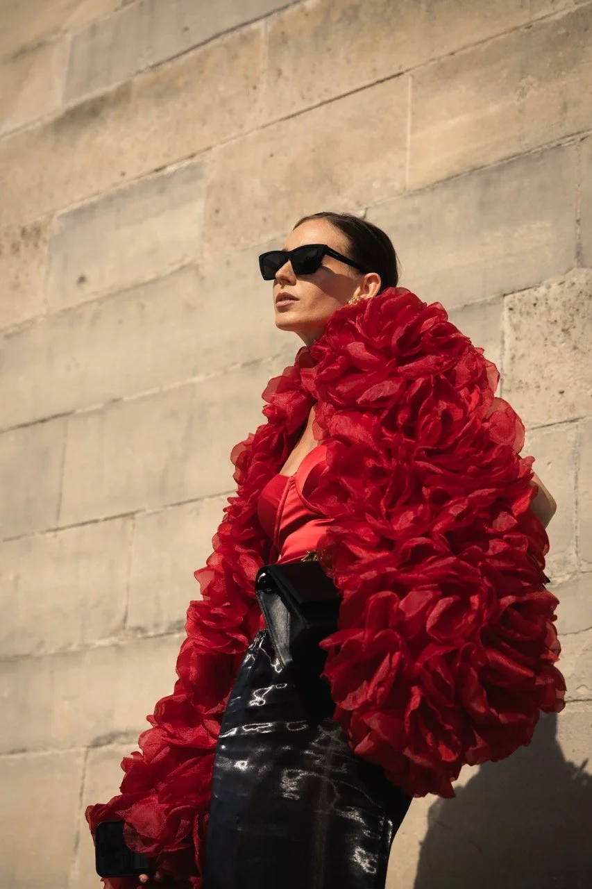 Eine modische Frau trägt eine auffällige rote Jacke mit großen Rüschen, schwarze Sonnenbrille, schwarze Lederhose und trägt eine schwarze Handtasche, posiert vor einer Steinfläche.