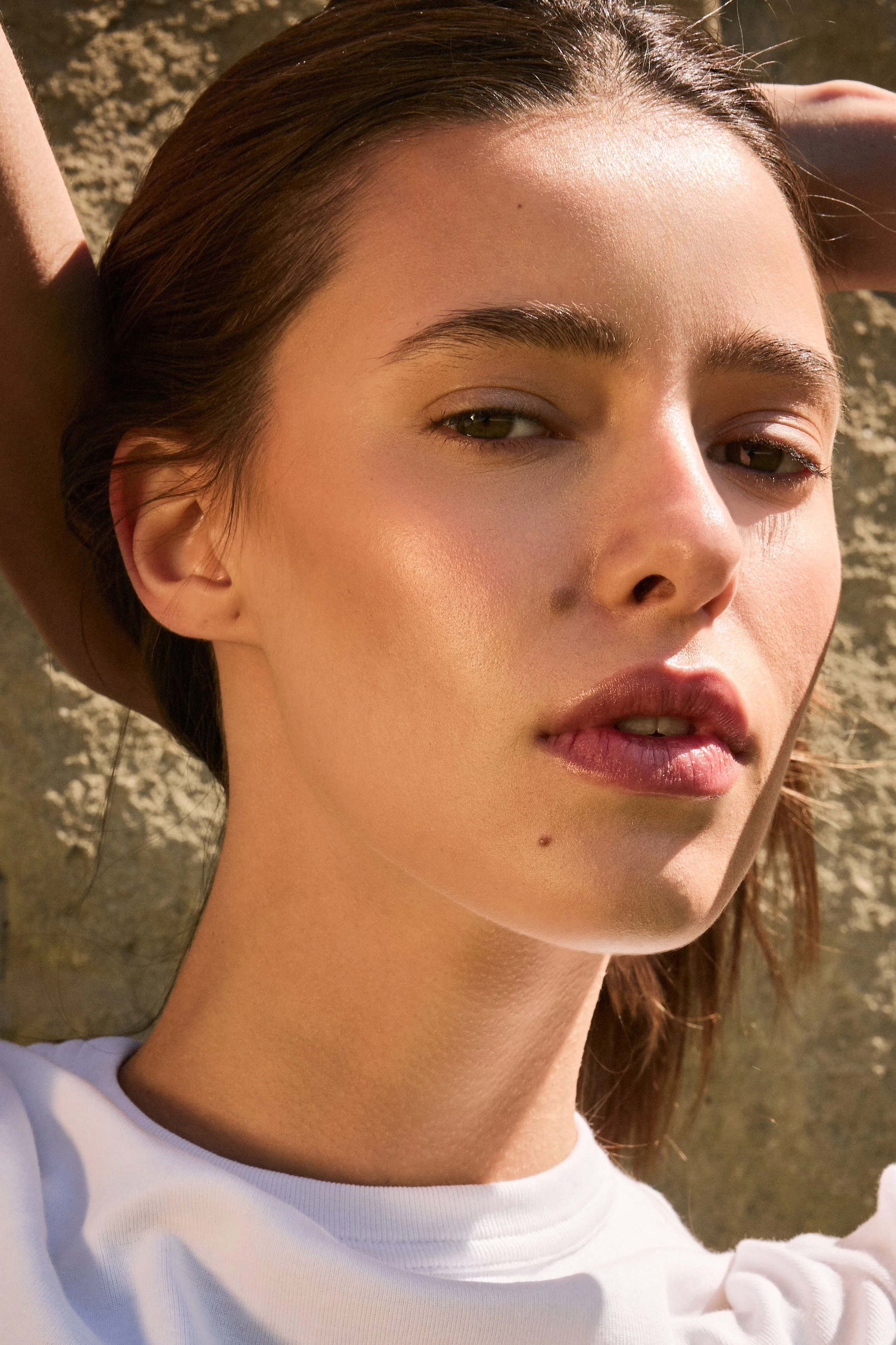 Close-up of a woman with brown hair lying on her back outdoors in sunlight, wearing a white shirt.