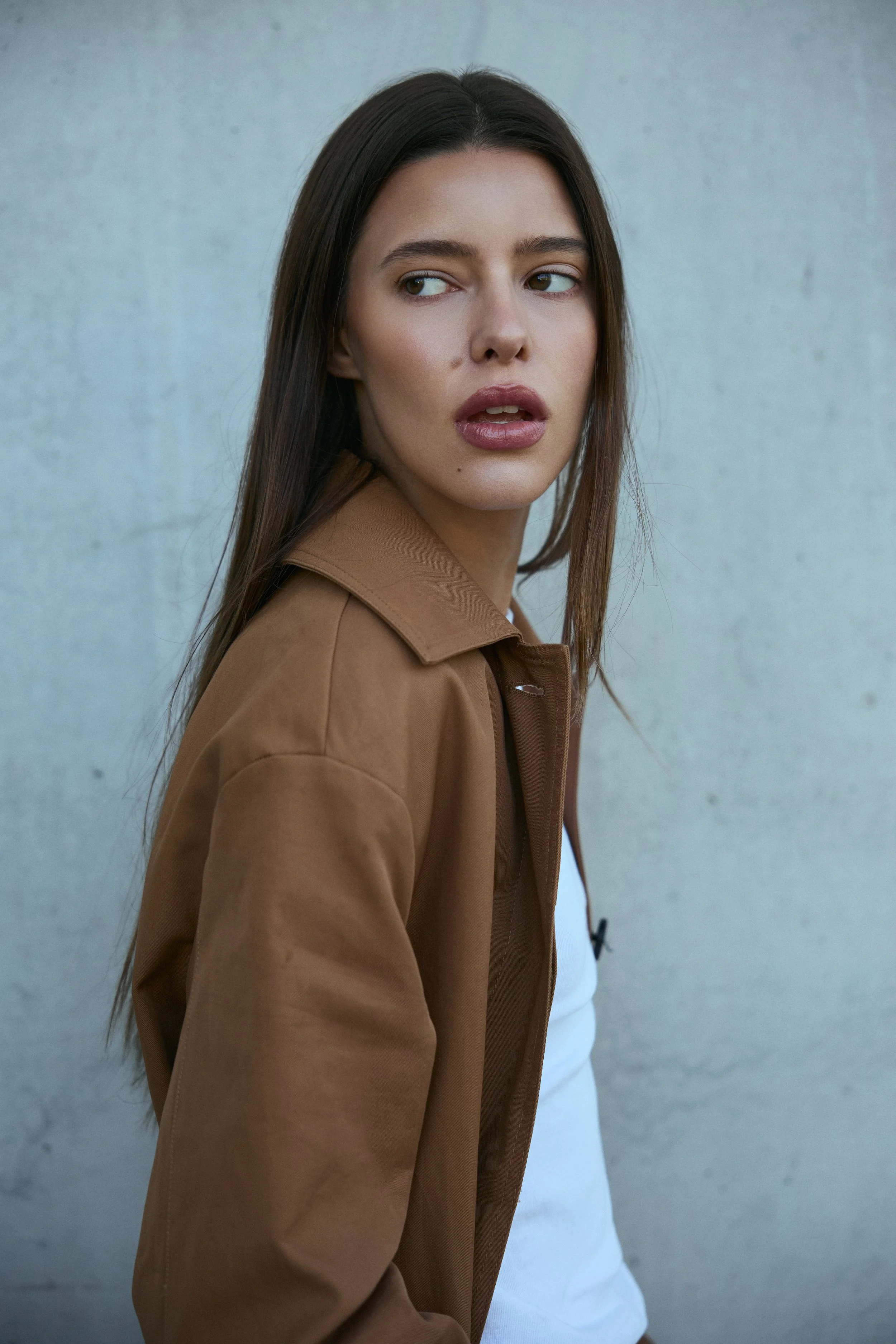 A young woman with long brown hair looking over her shoulder, wearing a brown jacket and white top standing against a plain gray wall.