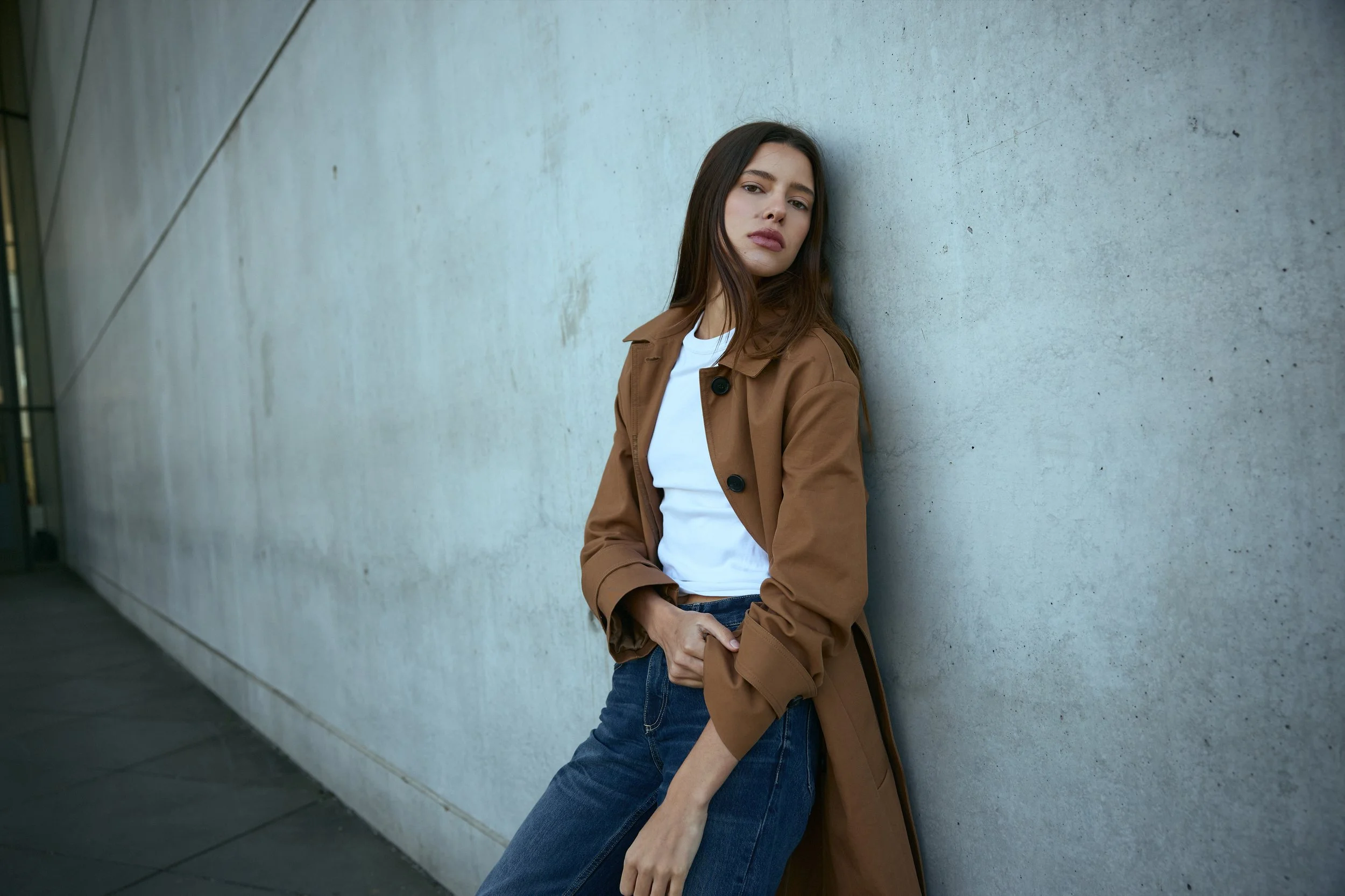 A woman with long brown hair leaning against a concrete wall, wearing a brown trench coat and blue jeans, looking at the camera with a neutral expression.