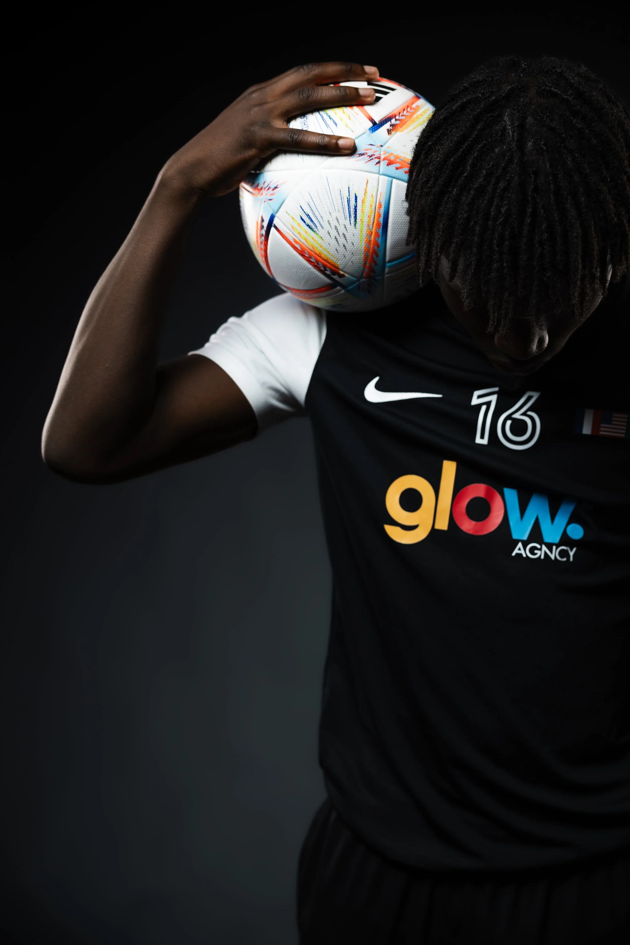 A soccer player holding a colorful Adidas UEFA Champions League soccer ball on his shoulder, wearing a black and white jersey with the number 16 and the word 'glow' on it, against a black background.