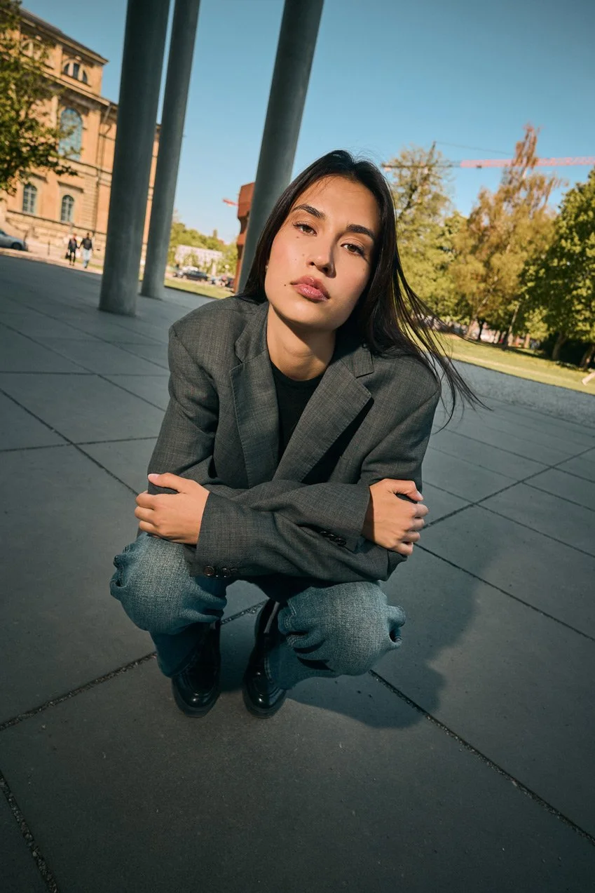 Young woman crouching outside on a paved surface, wearing a gray blazer and jeans, with a cityscape and trees in the background.