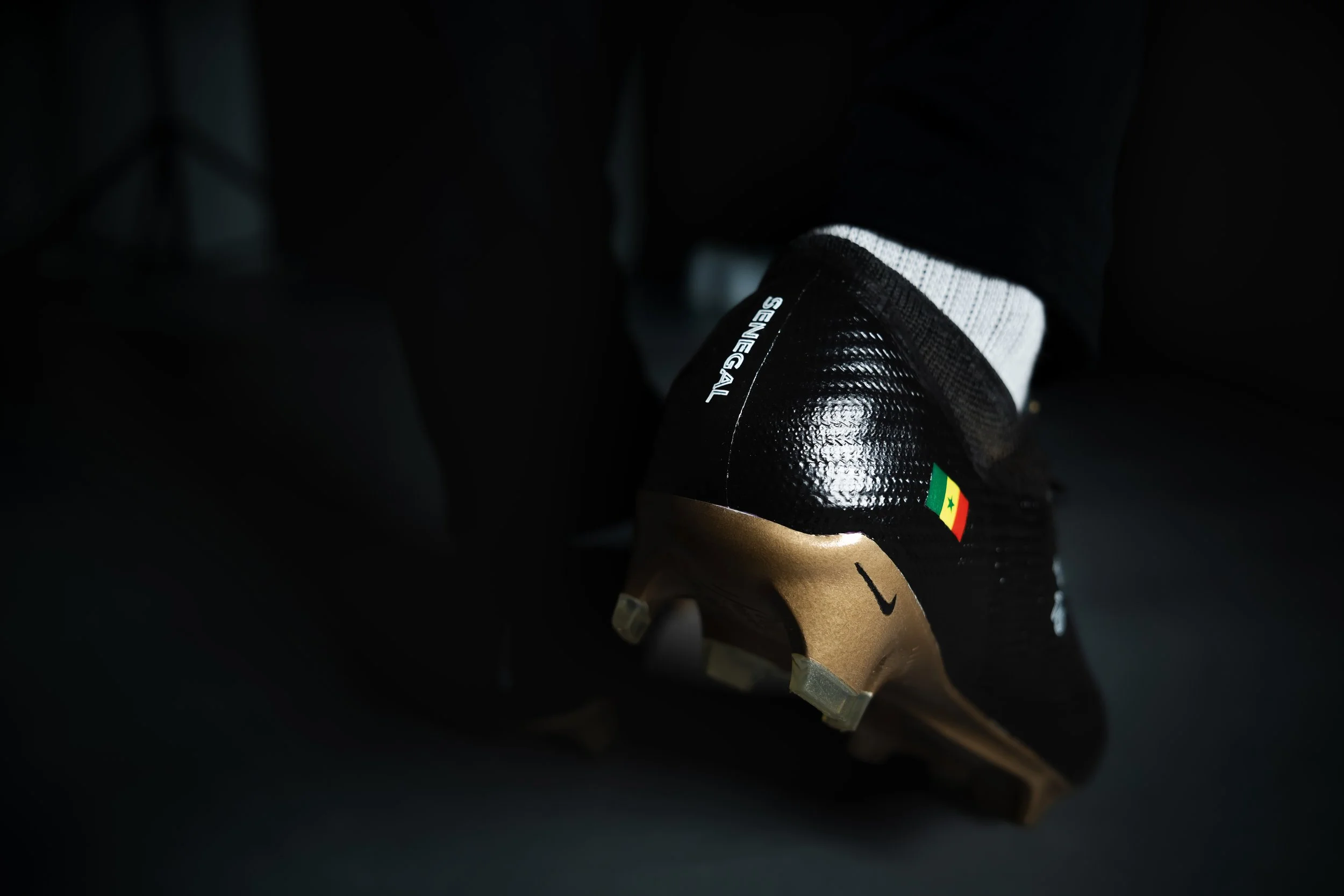 Close-up of a black soccer cleat with gold accents, featuring the Senegal flag and black laces, worn with black socks and black pants, on a dark background.