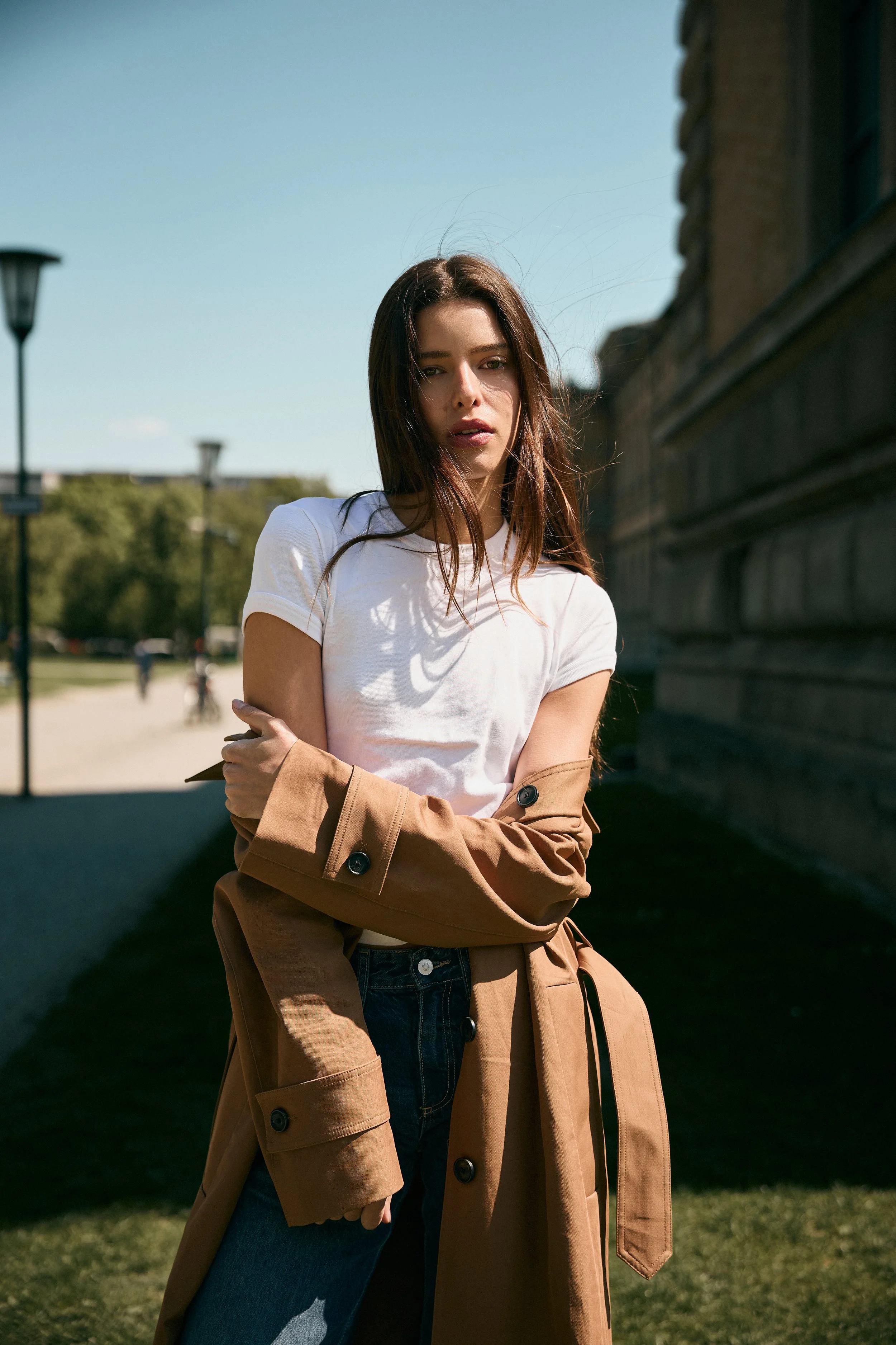 A young woman with long brown hair wearing a white t-shirt and blue jeans, standing outdoors near a stone wall on a sunny day. She has a brown trench coat draped over her shoulders and appears to be crossing her arms, looking at the camera.