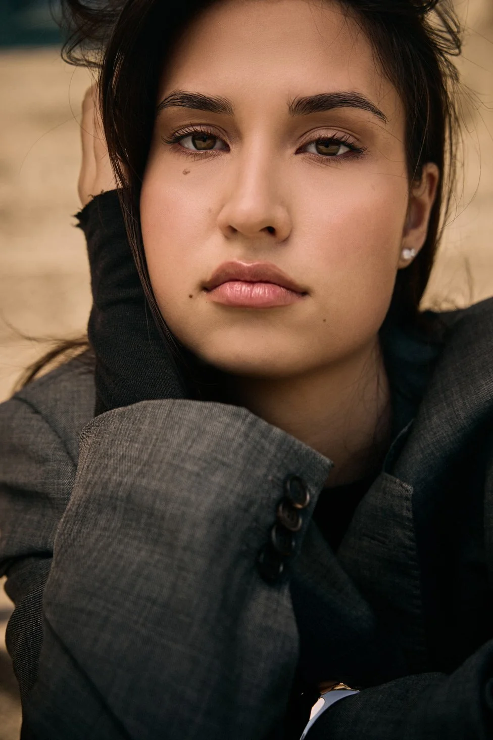 Close-up portrait of a young woman with dark hair, gray eyes, and light skin, wearing a gray blazer and black glove, outdoors with a blurred background.