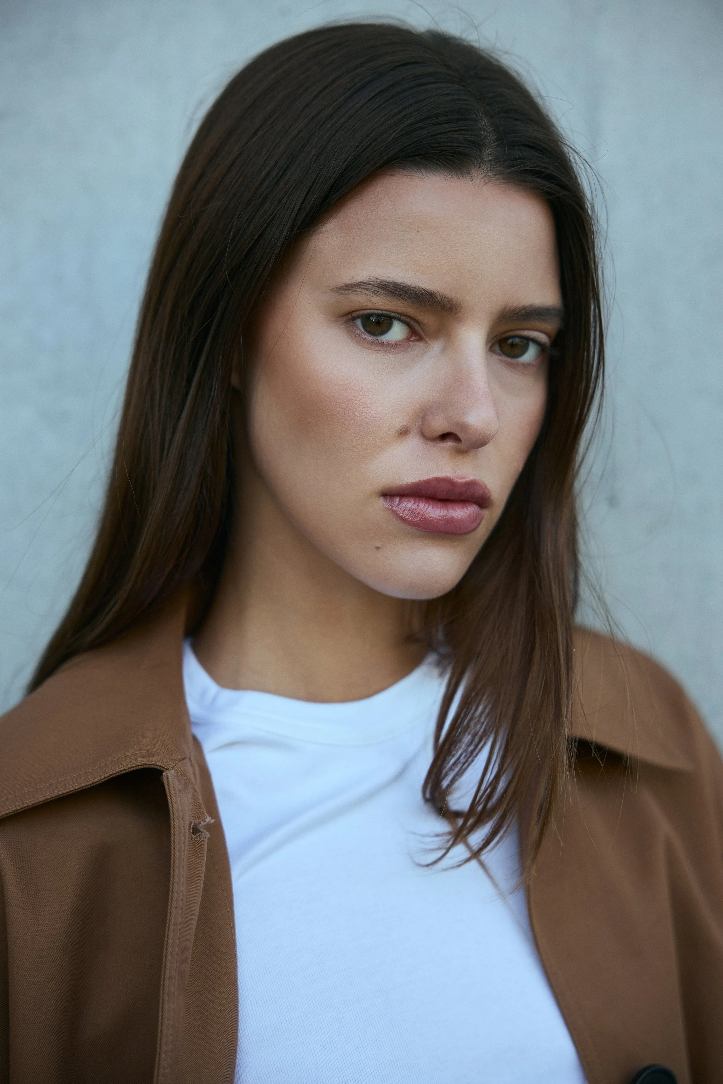 A young woman with brown hair, fair skin, and light makeup, wearing a white shirt and a brown jacket, standing against a gray background.