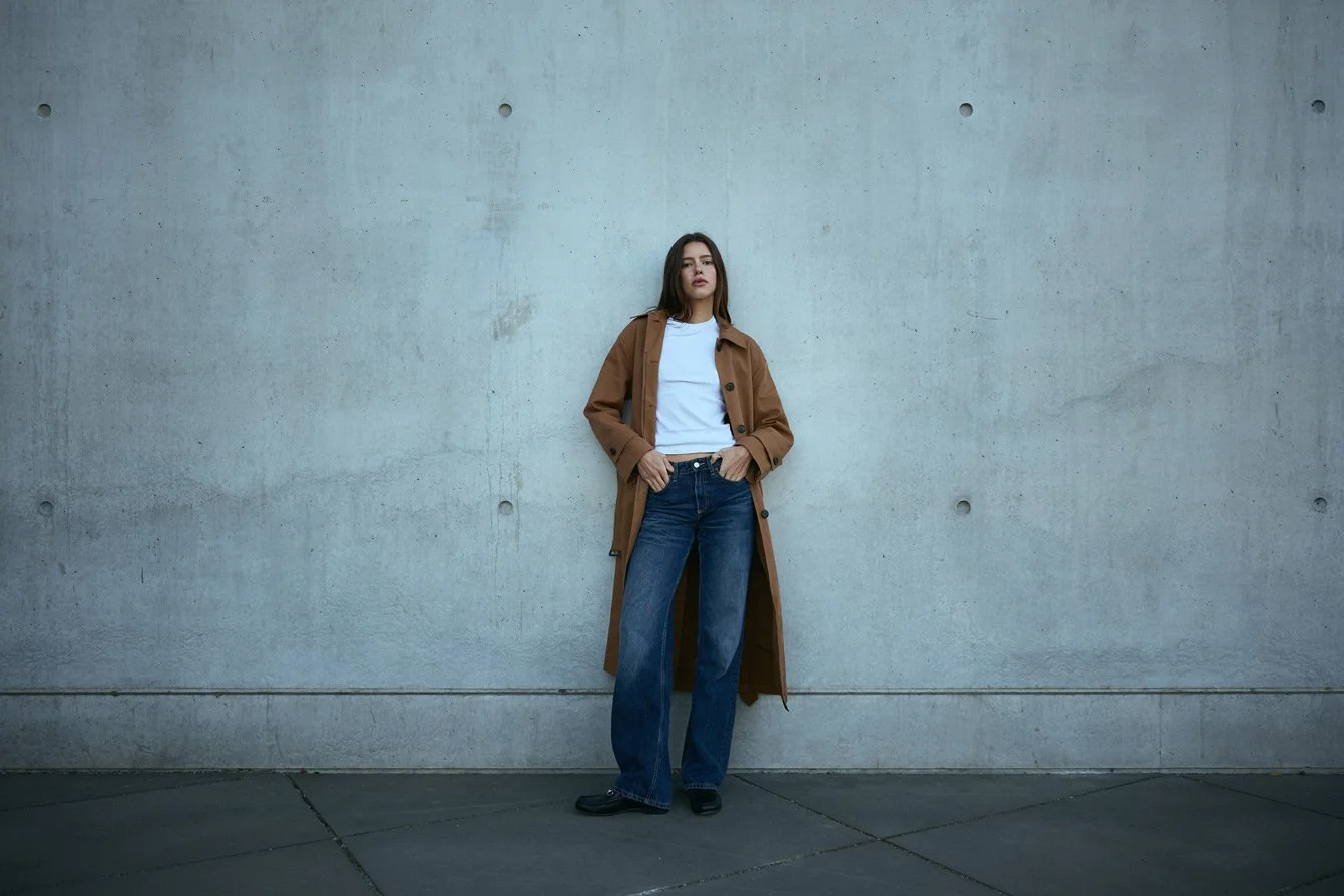Woman standing against a concrete wall, wearing a brown trench coat, white shirt, and blue jeans.