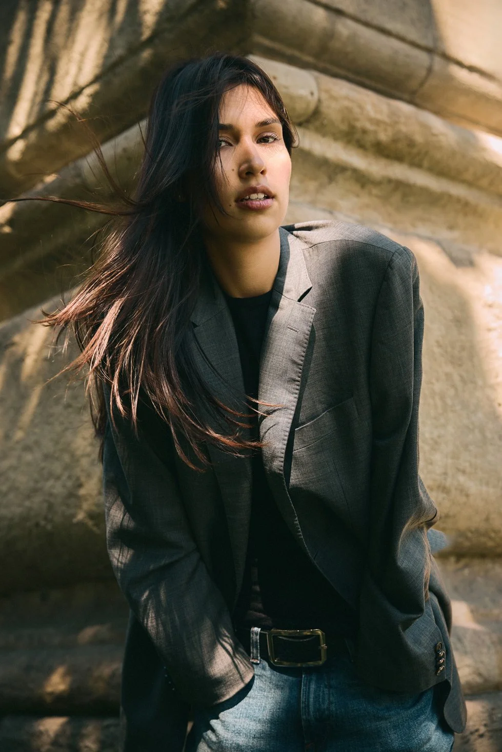A young woman with long dark hair, wearing a gray blazer and jeans, standing outdoors against a stone wall.
