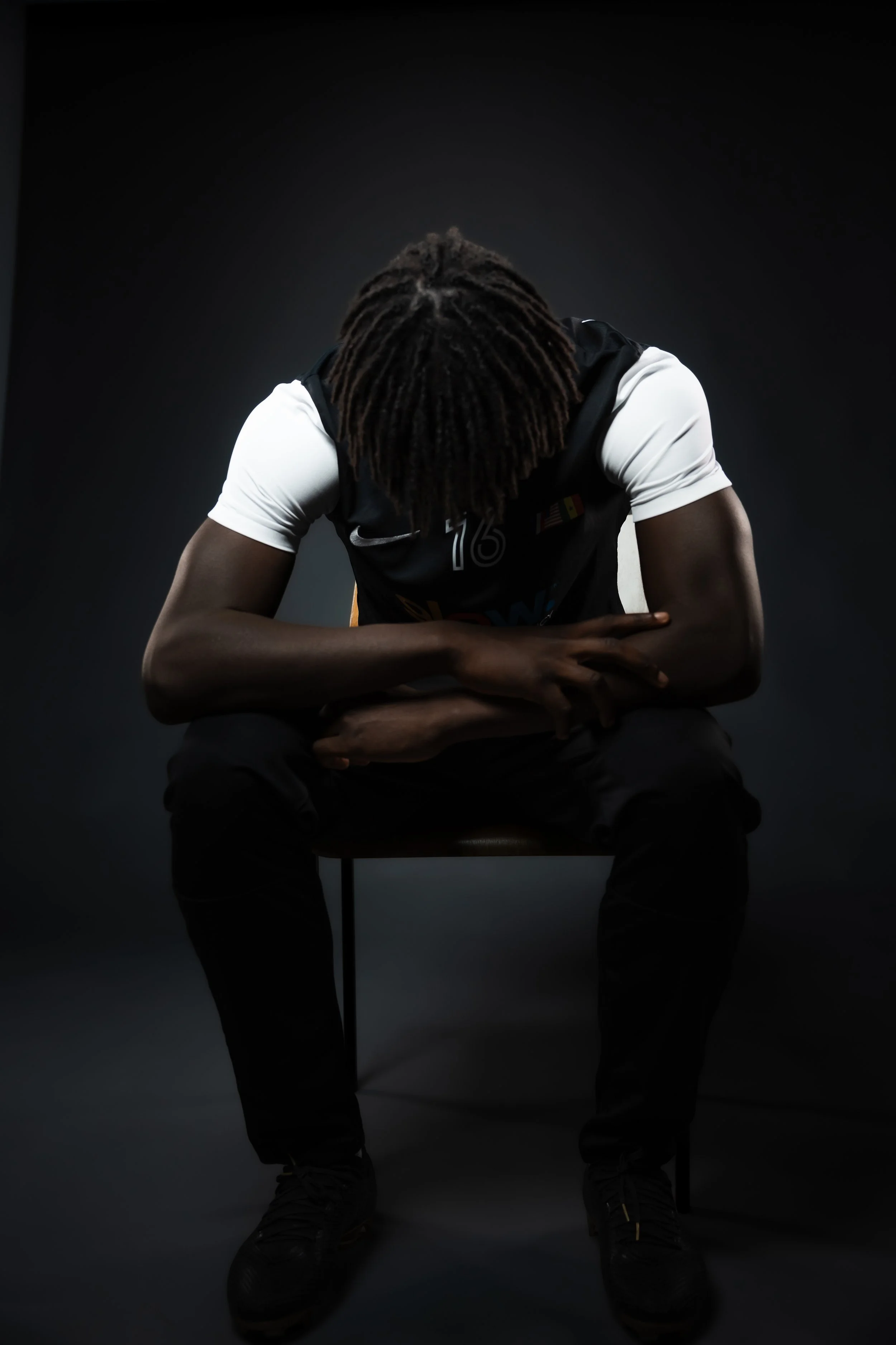 Person with dreadlocks sitting on a chair with head bowed and arms crossed, dressed in a black and white sports jersey, in a dark studio setting.