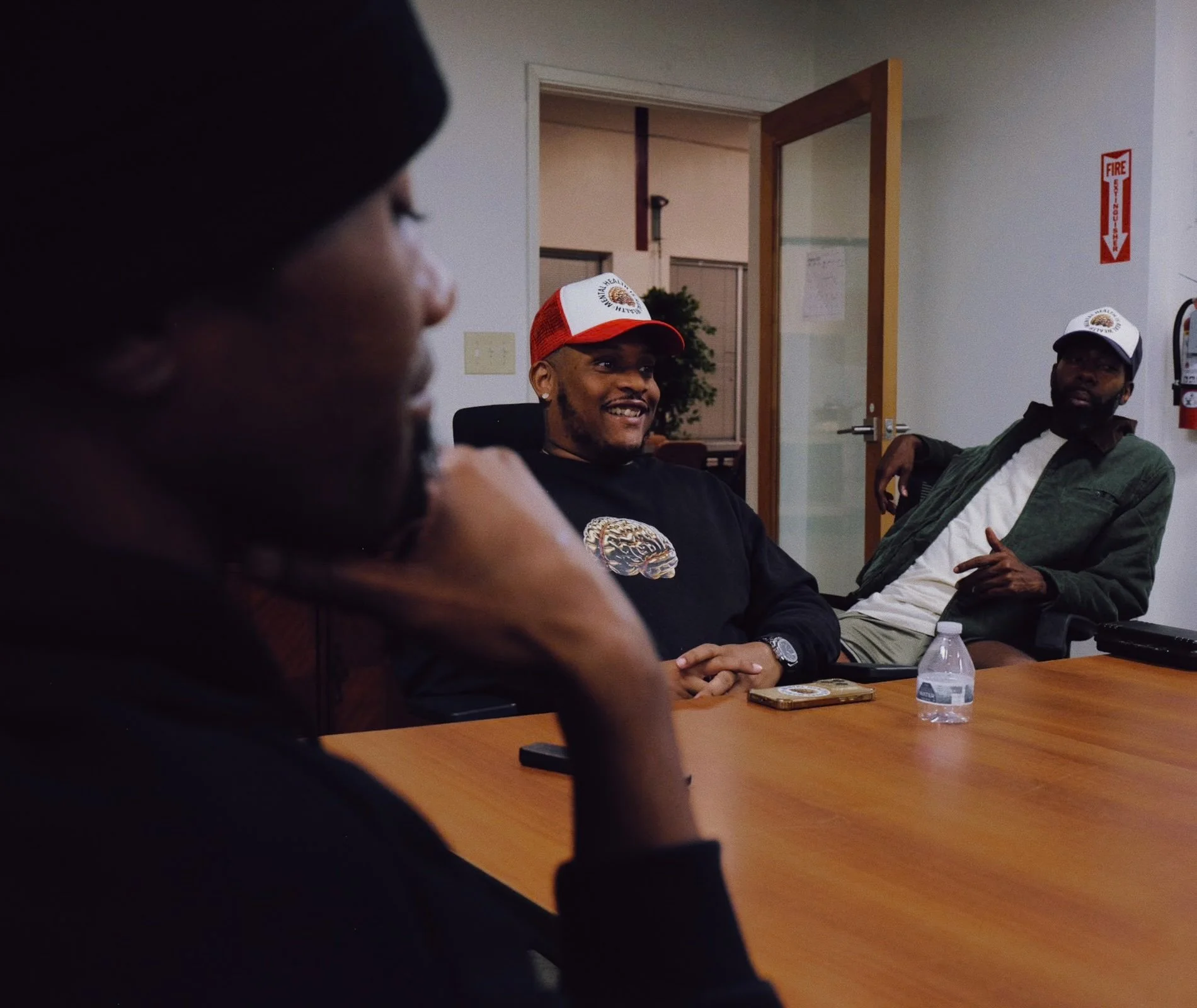 Three men sitting at a conference table, wearing hats with a fire department logo, in a room with a door, a fire alarm, and a water bottle on the table. One man is smiling, another is looking away, and the third is partially visible in the foreground.