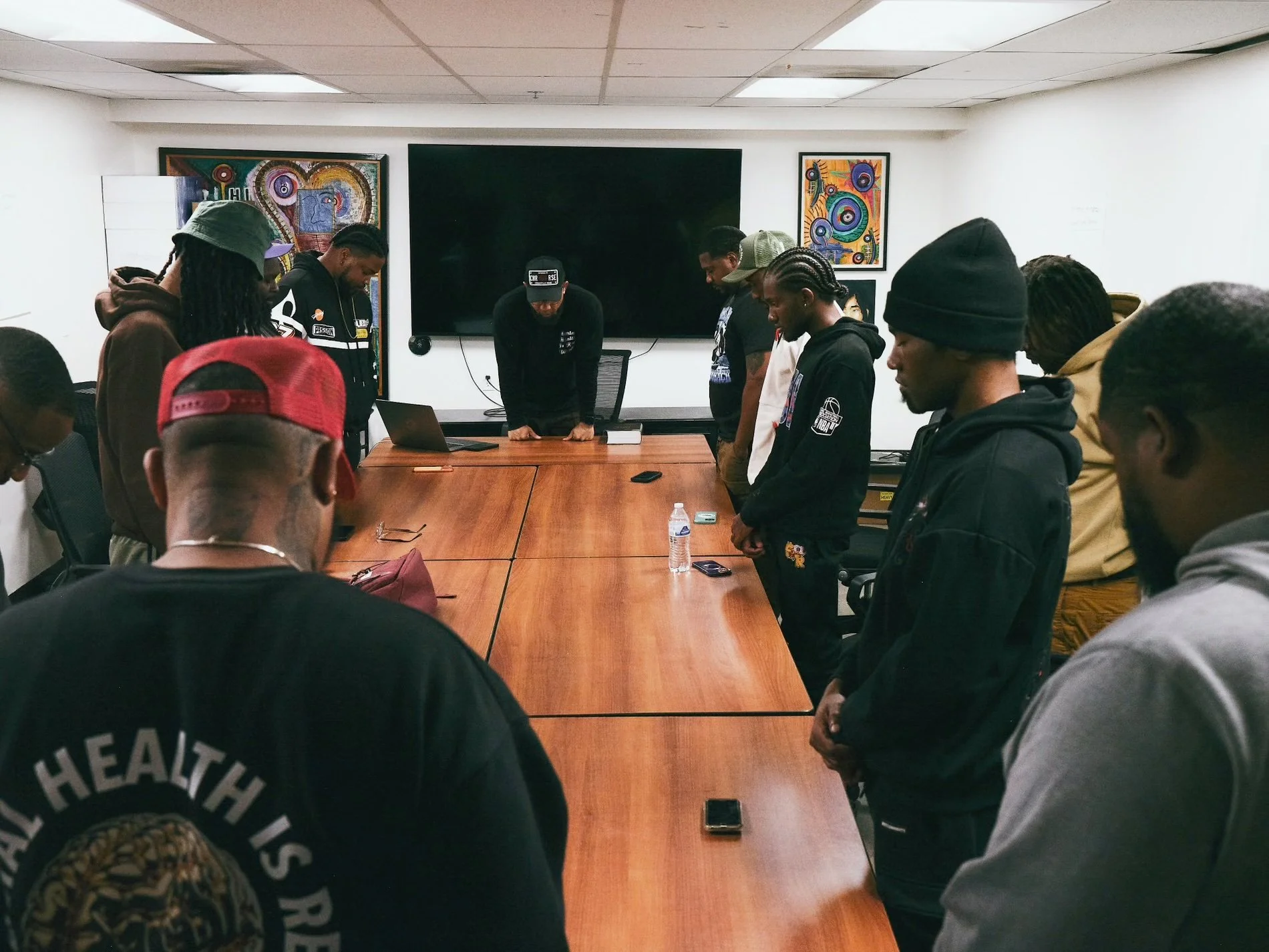 A group of people standing in a circle with their heads bowed in a meeting room with artwork on the walls and a large black screen on the wall.